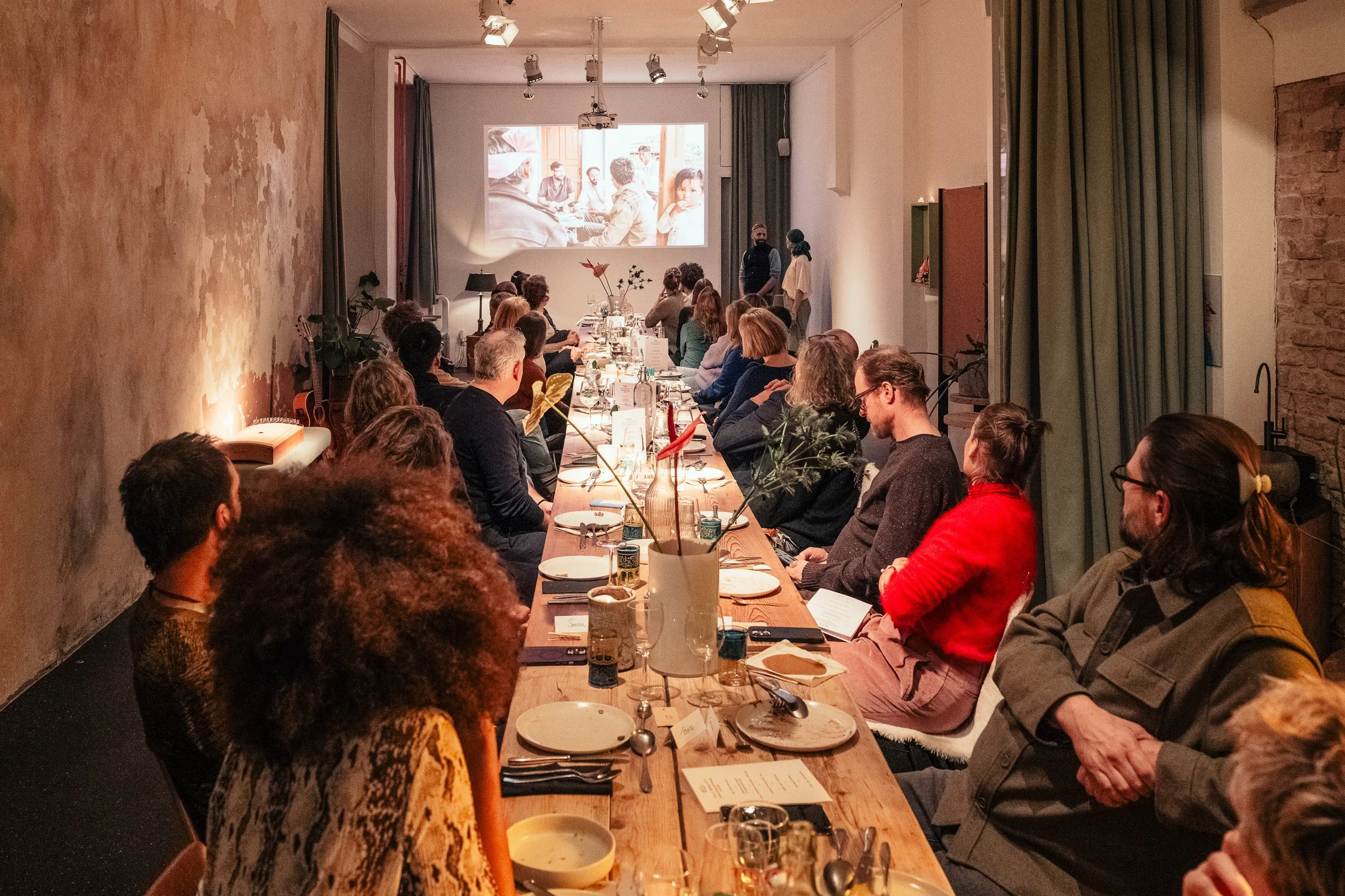 Ein Raum mit warmem Licht am Abend. Eine lange gedekte Tafel mit Menschen die auf eine Wand mit einer Videoprojektion sehen.