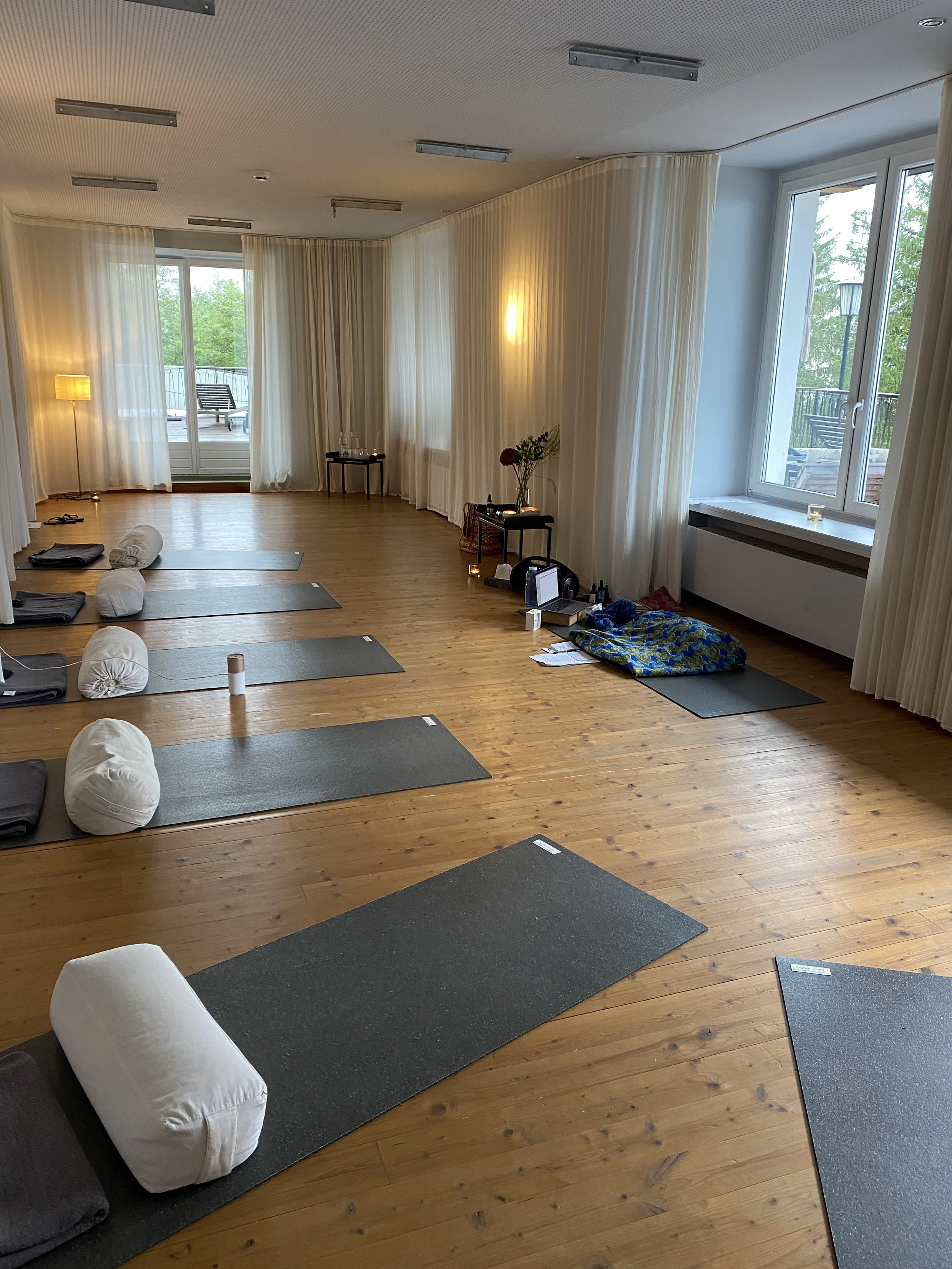 Ein ruhiger Raum mit Yoga-Matten, Kissen, Decken und einem Tee, vorbereitet für eine Yoga- oder Meditationsklasse. Große Fenster lassen viel natürliches Licht herein und zeigen eine grüne Umgebung draußen.