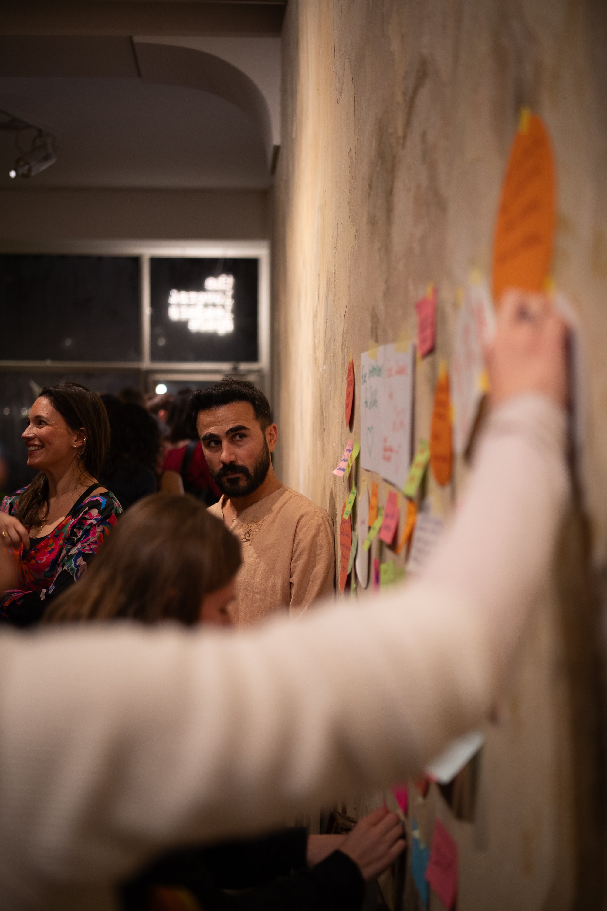 Menschen in einer gemütlichen, hell erleuchteten Räumlichkeit, einige hängen an eine Wand mit bunten Post-its, während andere zuschauen oder sprechen.