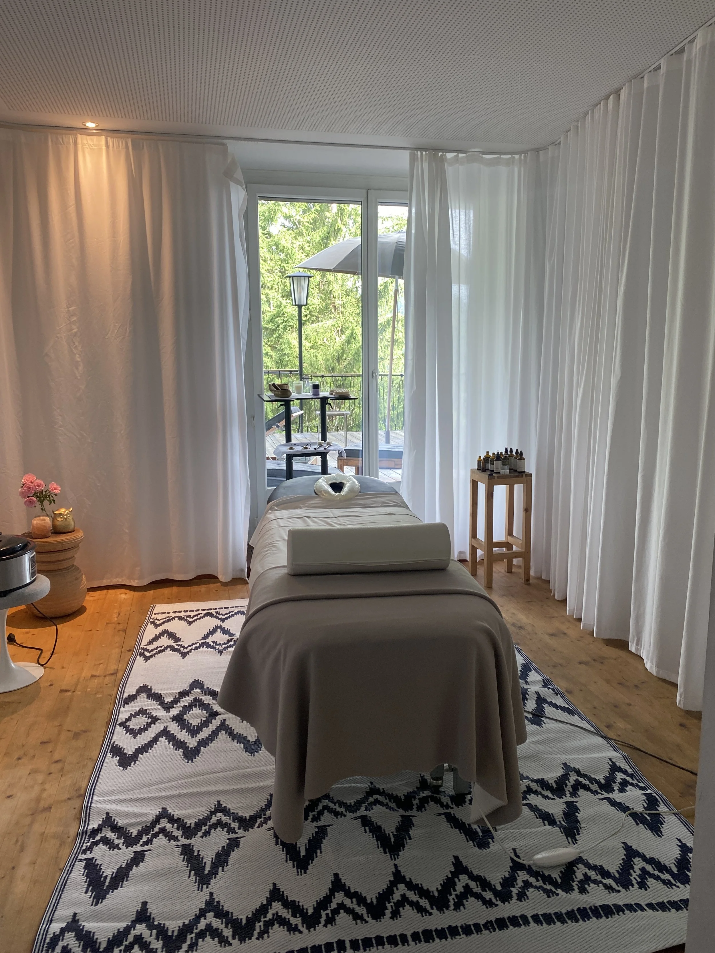 Ein Behandlungsraum mit einem Massagebett, weißen Vorhängen, einer kleinen Terrasse mit Stühlen und einem Sonnenschirm, violette Blumen und verschiedene Flaschen auf einem kleinen Regal.