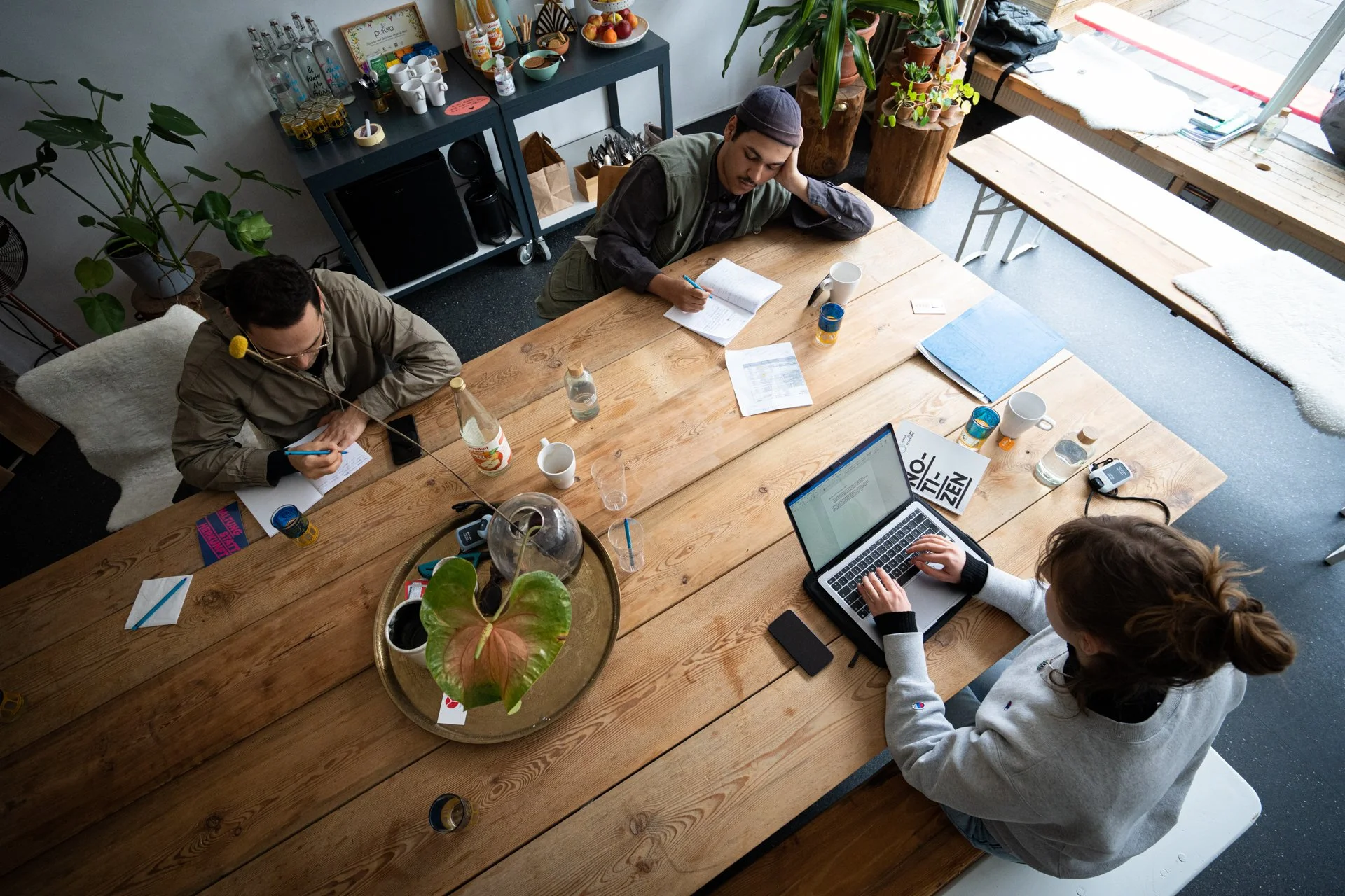 Ein gemütlicher Raum mit drei Menschen die an einem Holztisch  sitzen und mit Papier, Stift und Laptop arbeiten.