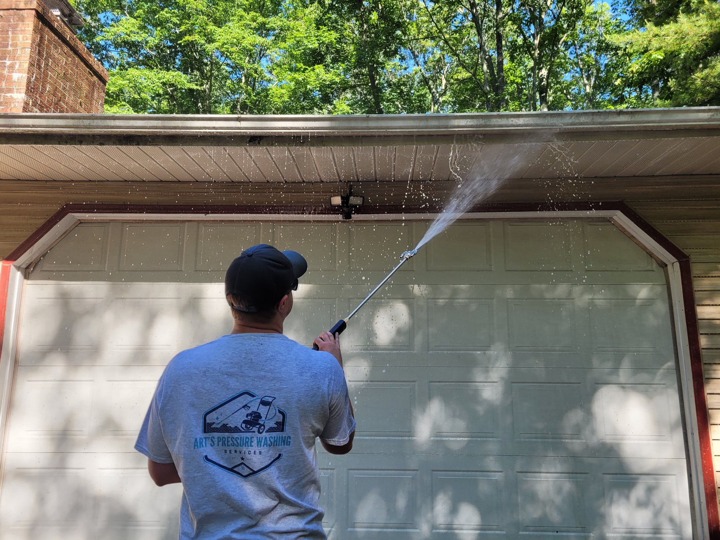 Person pressure washing a garage door under an overhang with trees and blue sky overhead.