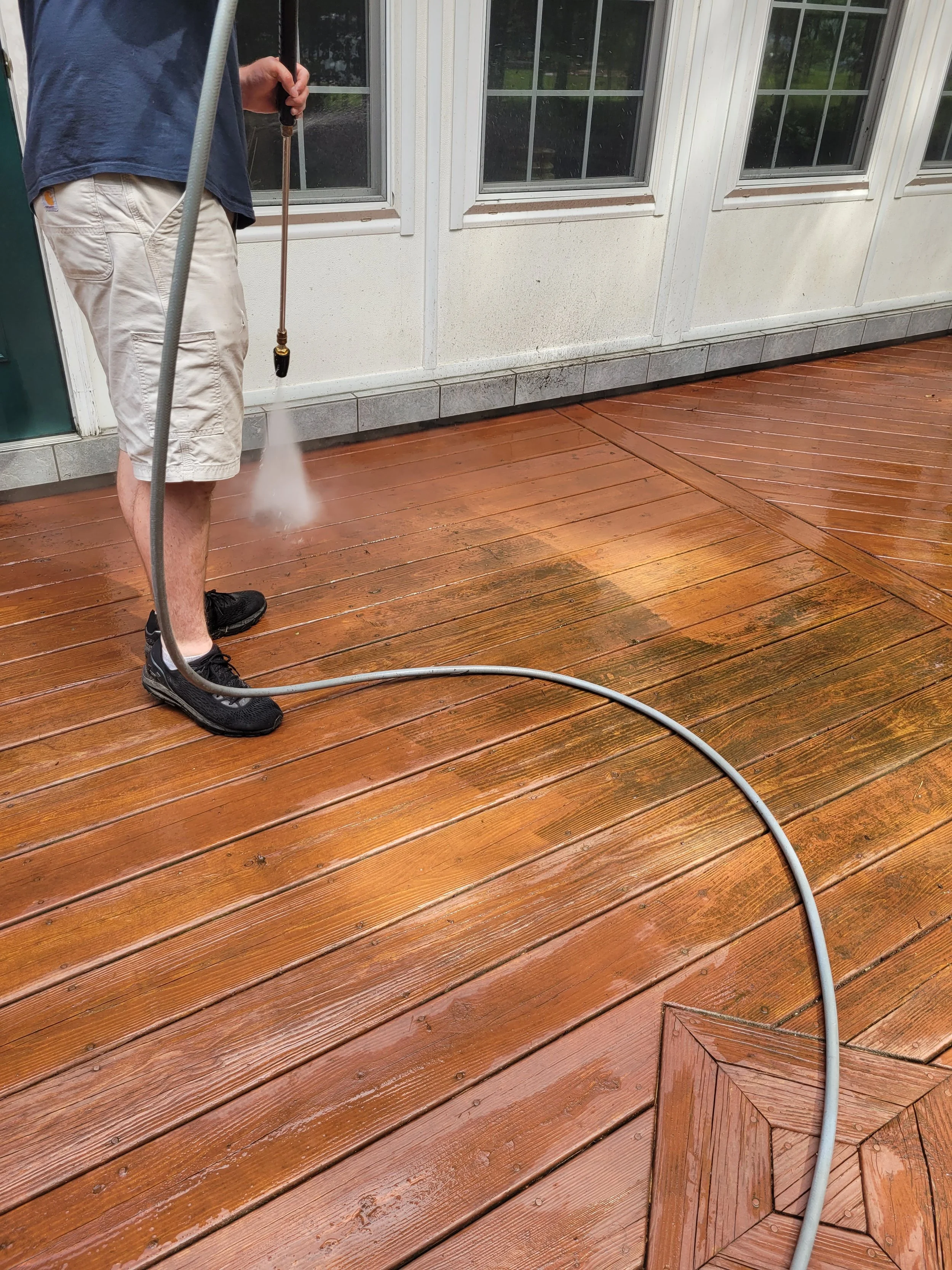 A person wearing black sneakers and beige shorts is pressure washing a wooden deck. The person is holding a water hose/spayer and spraying water on the deck, which appears wet. The deck is next to a house with white walls and large windows.
