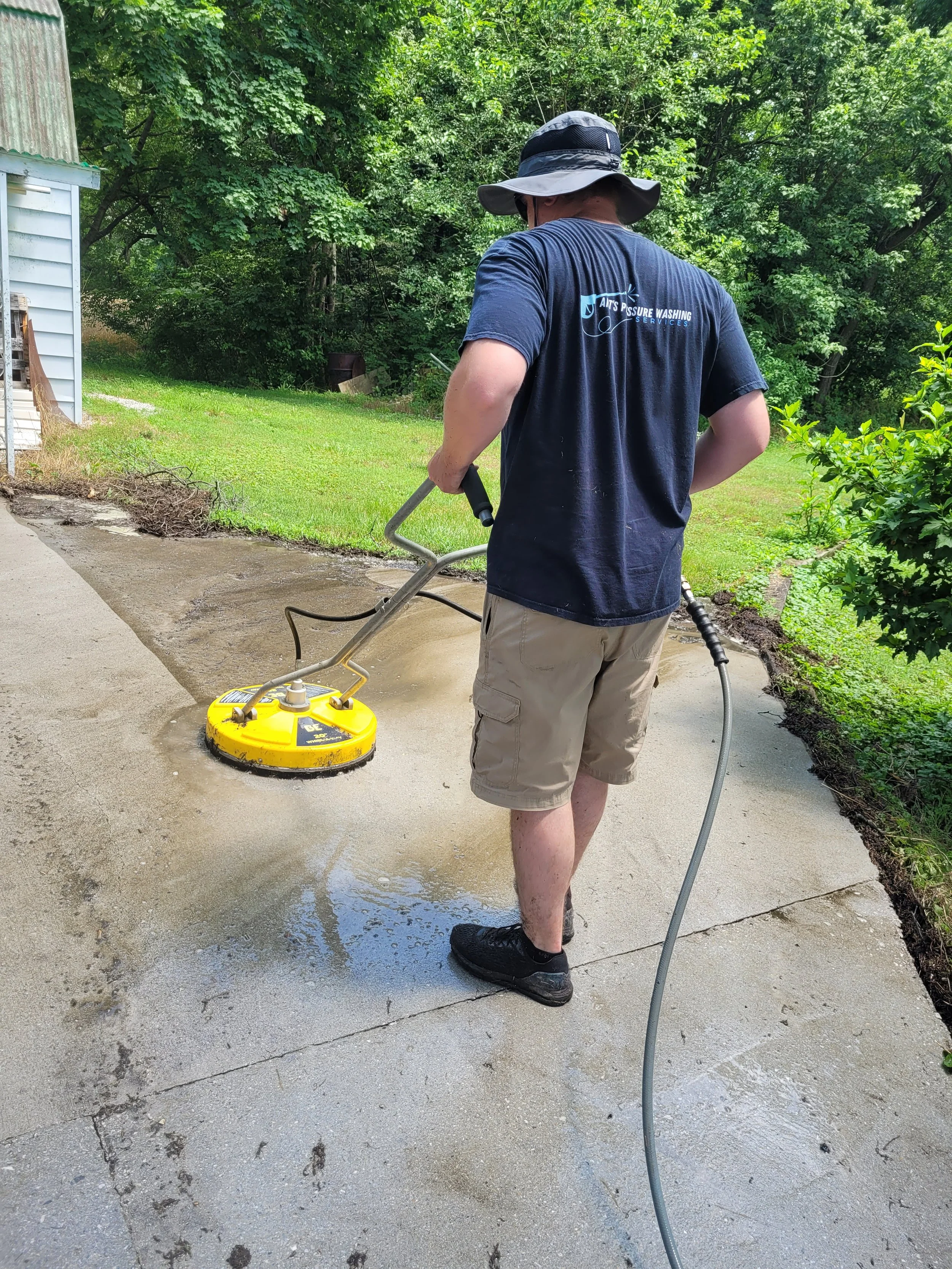 Man using a power washer to clean concrete sidewalk in a backyard, wearing a wide-brim hat, navy blue t-shirt, khaki shorts, and black shoes.