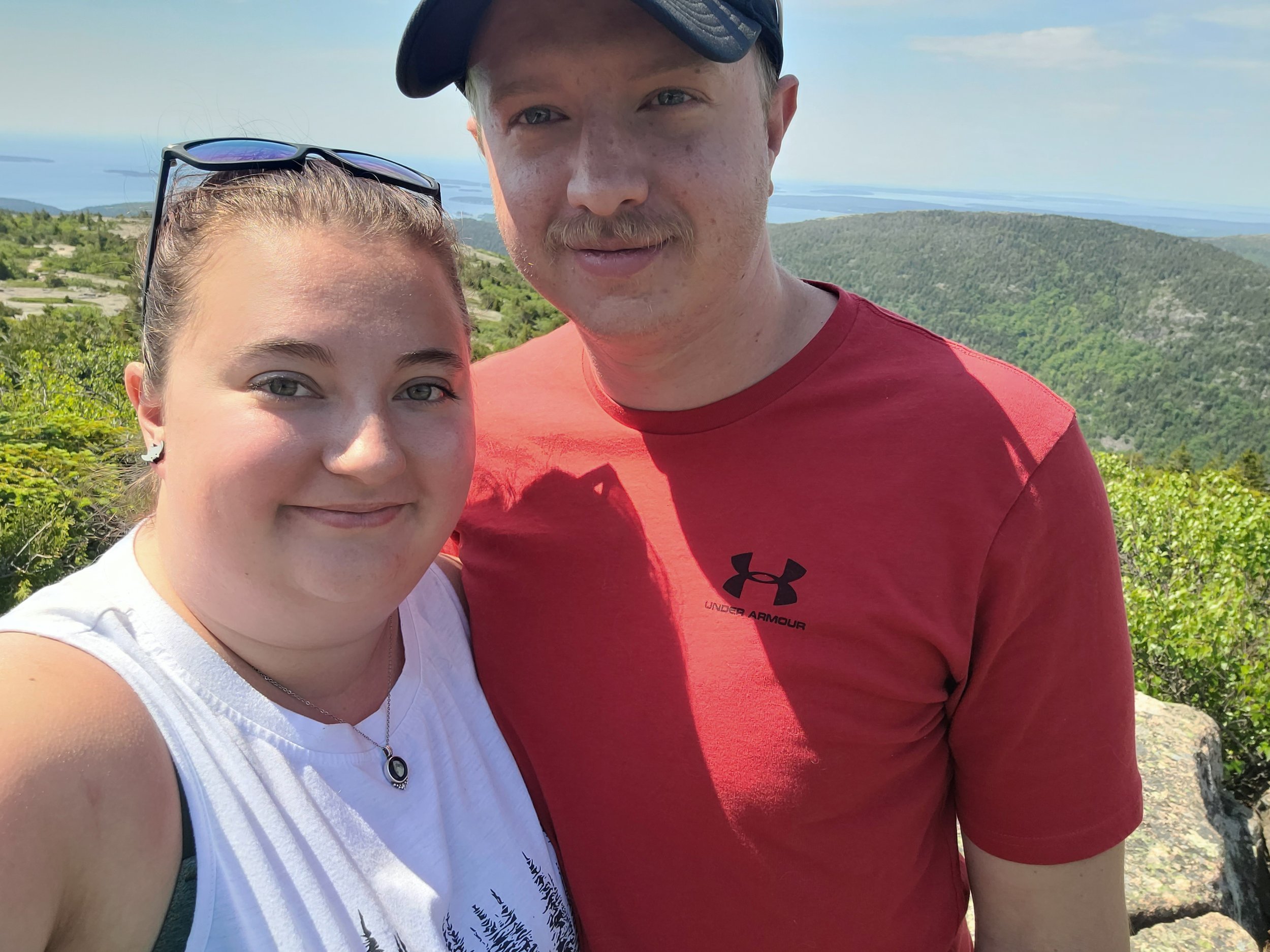 A young woman and a man are taking a selfie outdoors on a mountain overlooking a green landscape and a large body of water in the distance. The sky is clear and blue.