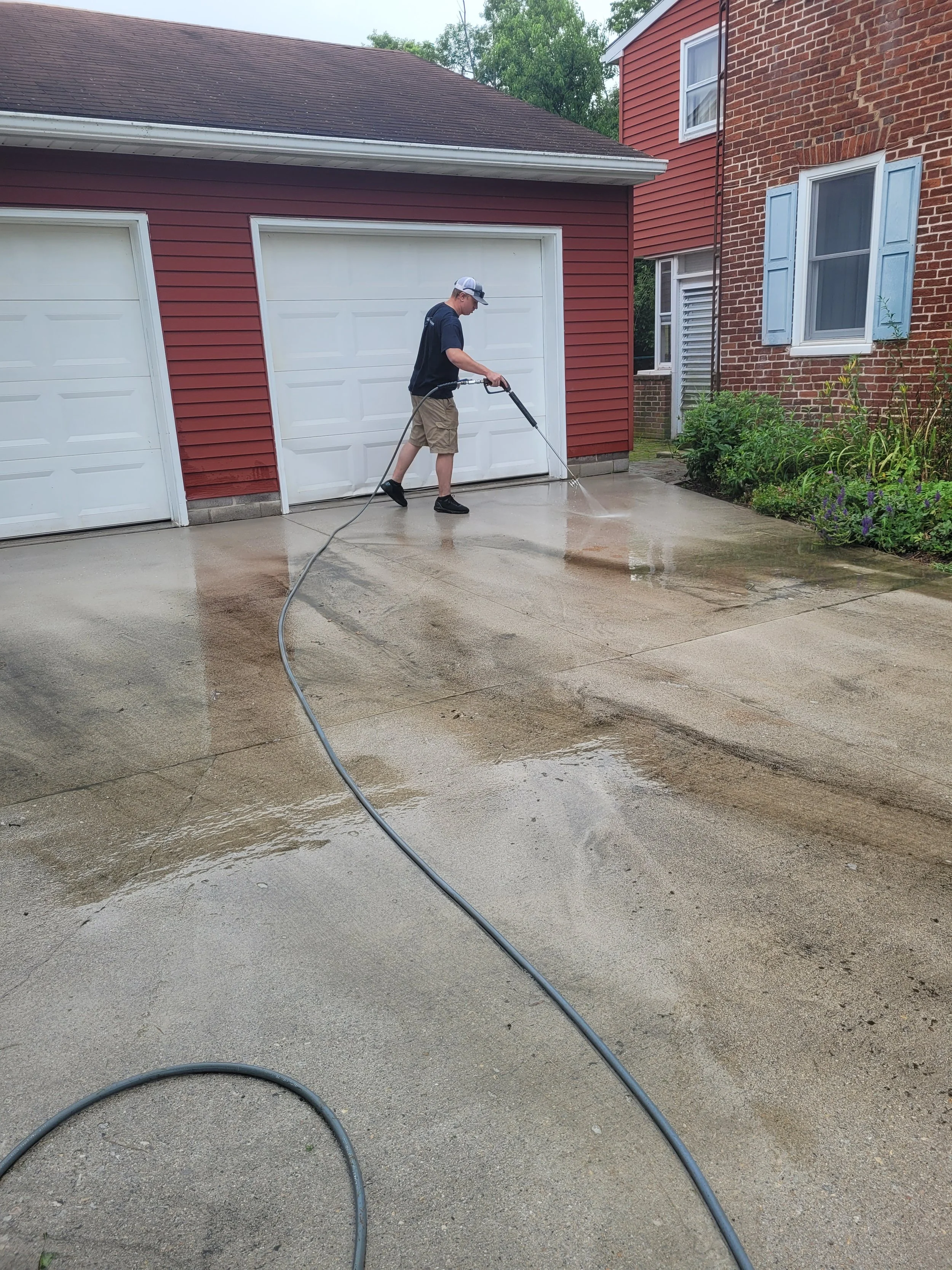 A man pressure washing a concrete driveway in front of a red garage and a brick house.