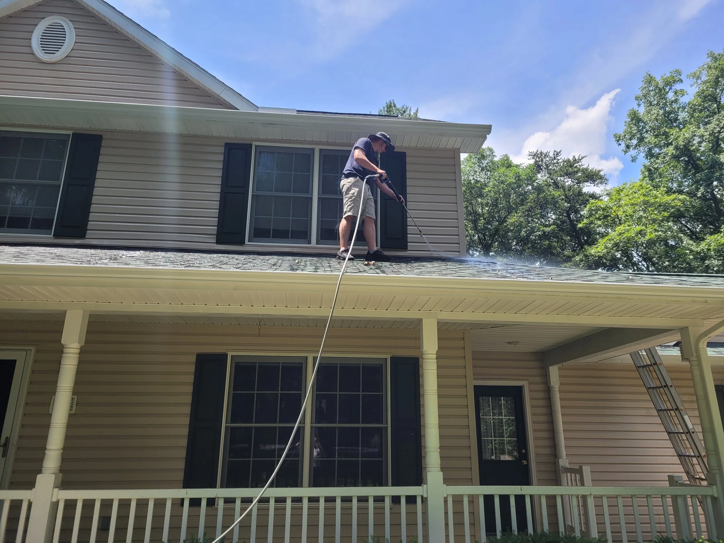 A man is cleaning the roof of a house with a power washer on a sunny day. There are trees and a clear blue sky in the background, and the house has beige siding and black shutters.