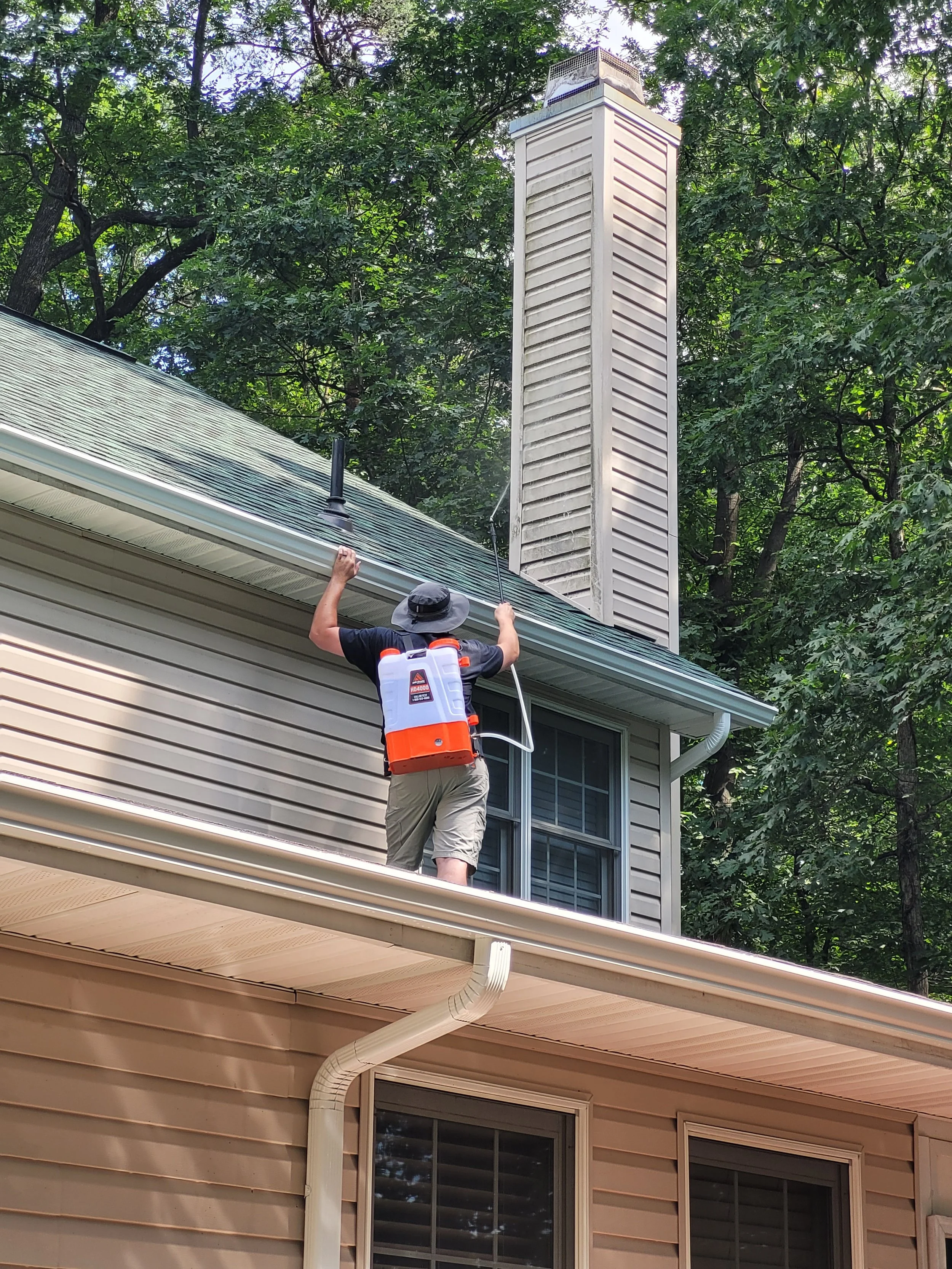 Person spraying pesticide on house roof with garden hose, wearing a hat and carrying a backpack sprayer