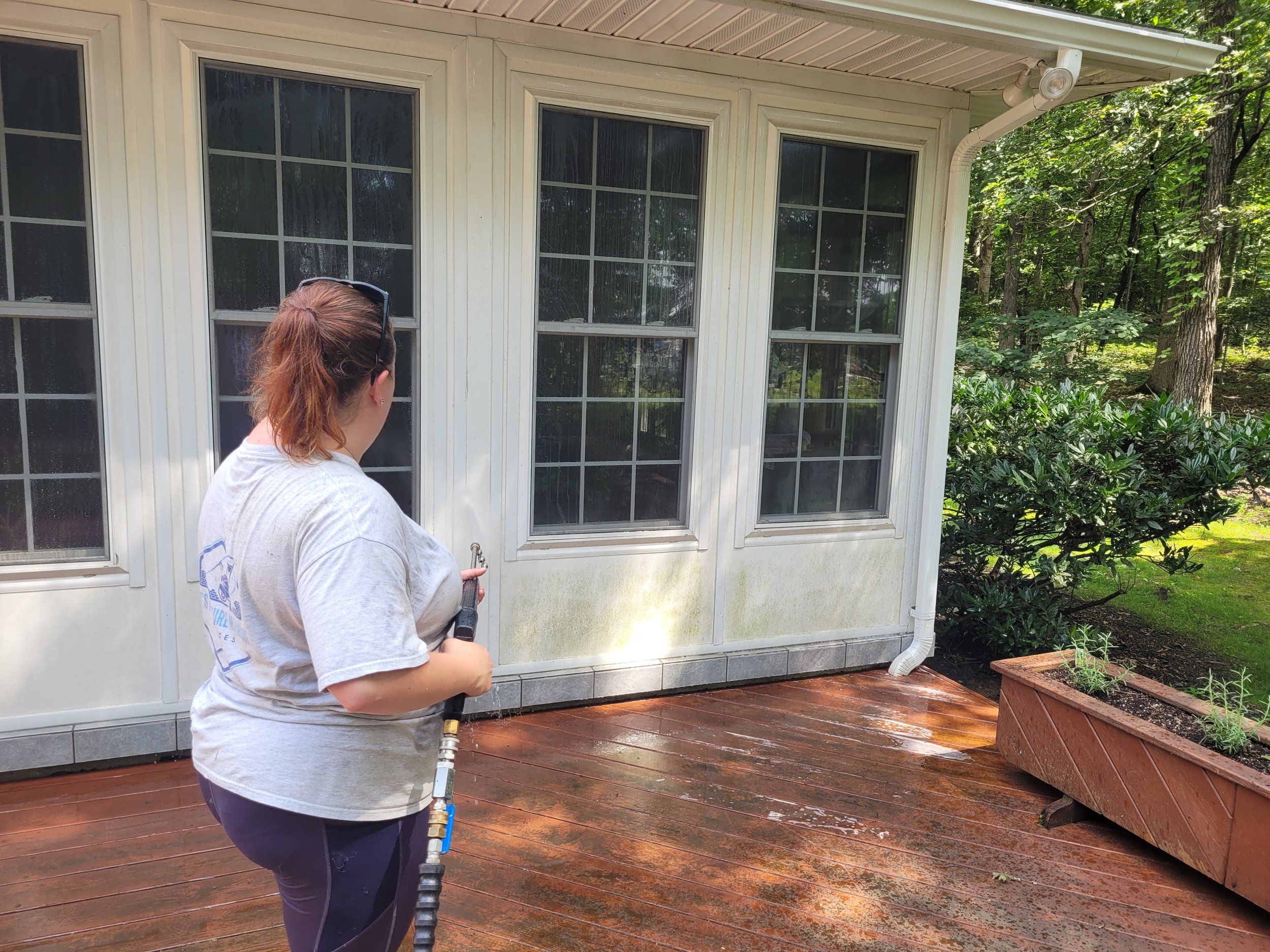 A woman with reddish hair, wearing a gray t-shirt and sunglasses on her head, stands on a wooden deck holding a hose and spraying water onto the exterior of a house with white walls and large windows. Surrounding greenery and trees are visible in the