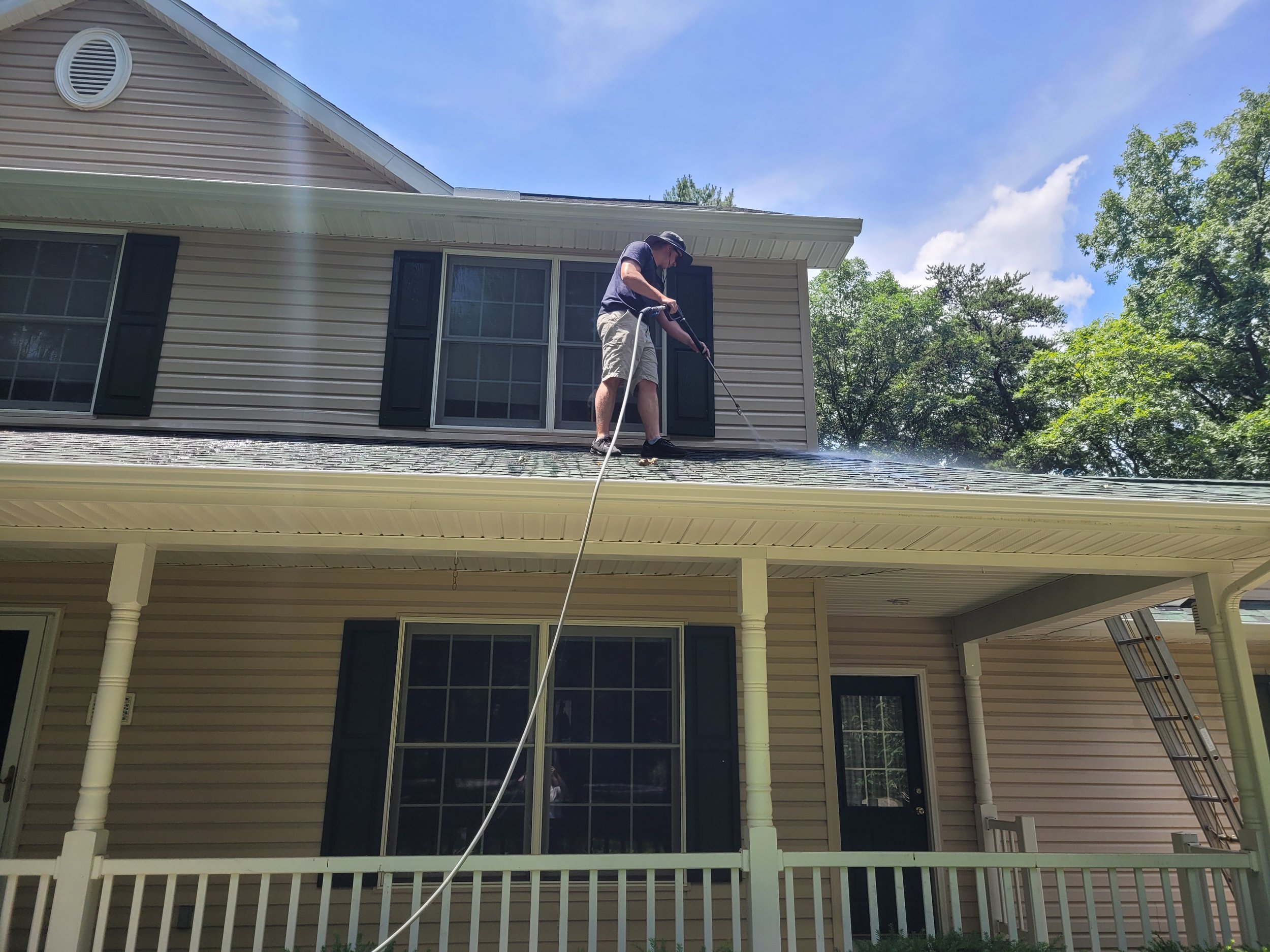 A man wearing a hat and shorts is standing on the roof of a house, using a pressure washer to clean the roof.