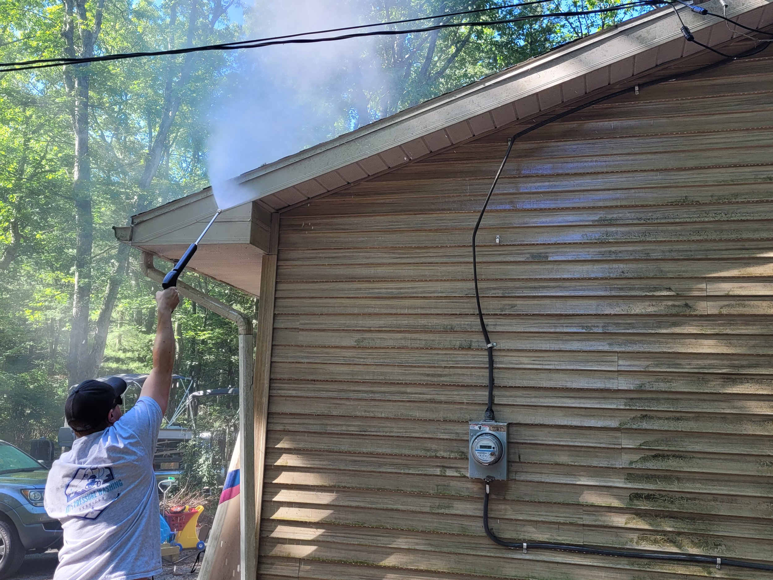 A person using a pressure washer to clean the exterior of a wooden house's wall, with green trees and blue sky in the background.