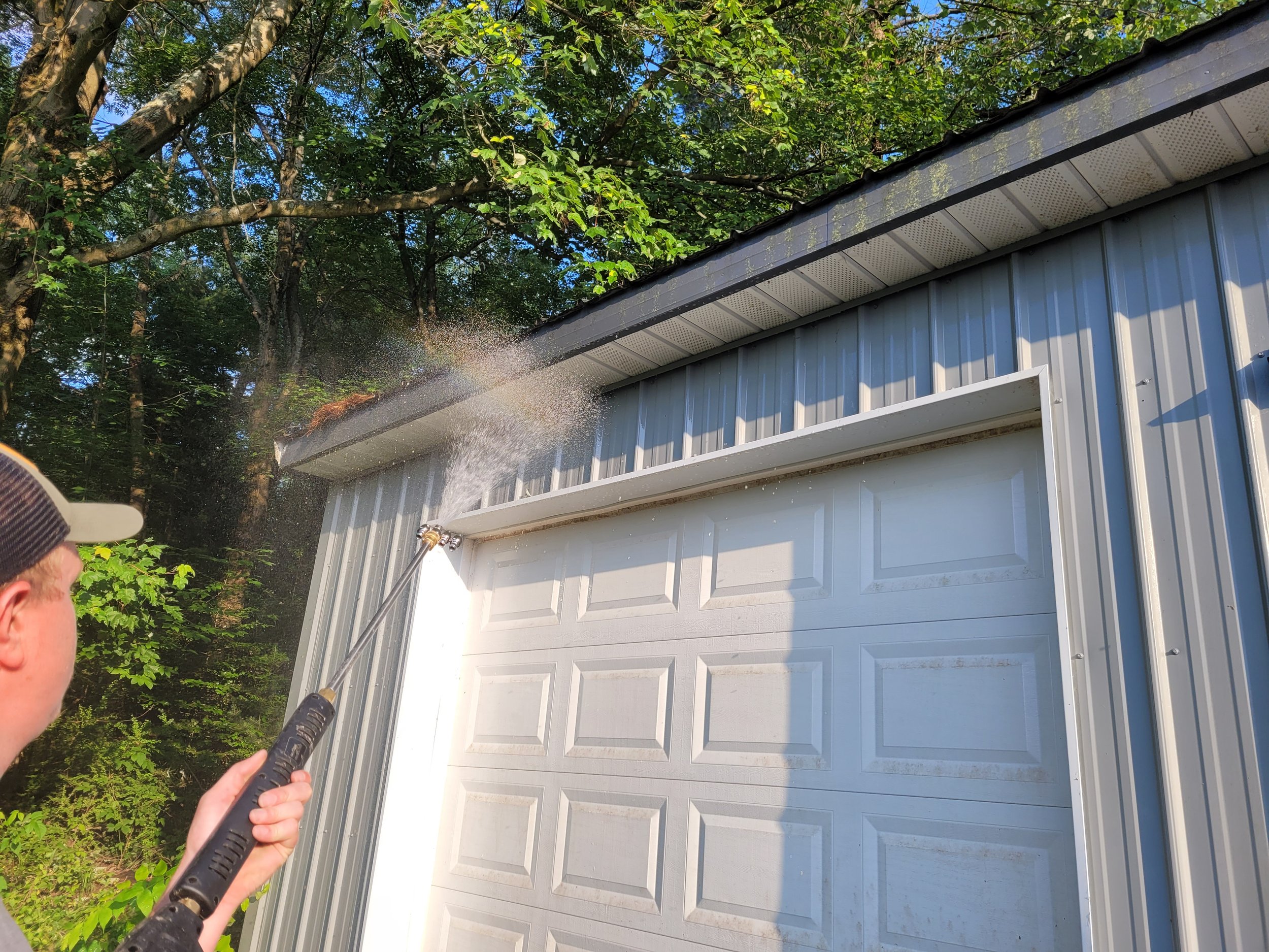 A person pressure washing a white garage door with a garden hose, with trees and blue sky in the background.