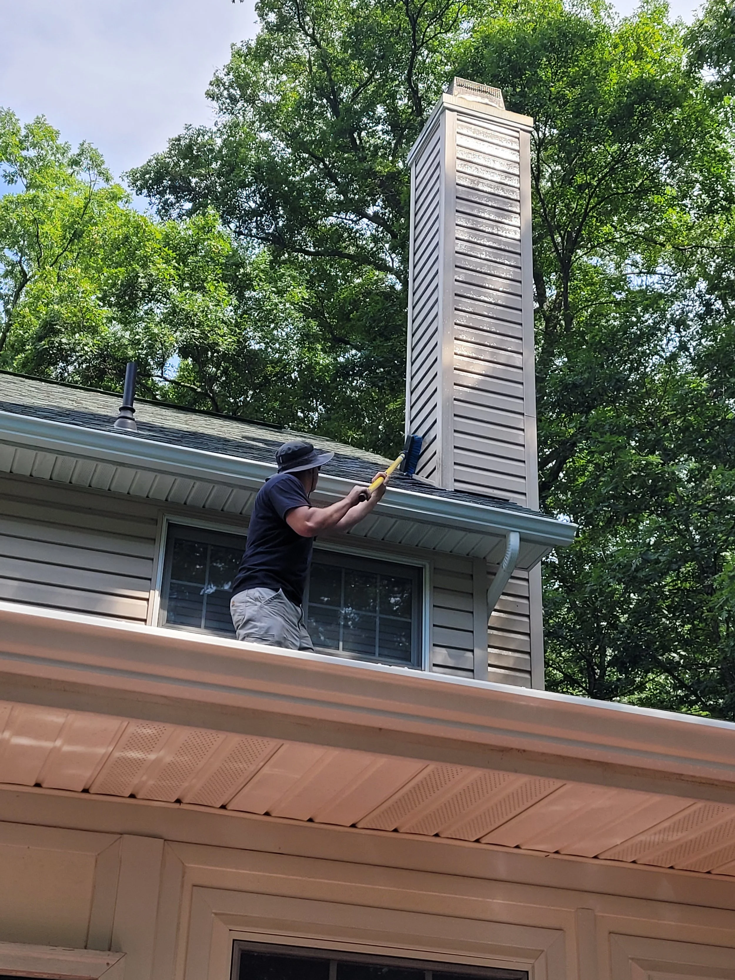 A person wearing a wide-brimmed hat is standing on a house's balcony, cleaning the chimney through an opening in the roof, with lush green trees in the background.