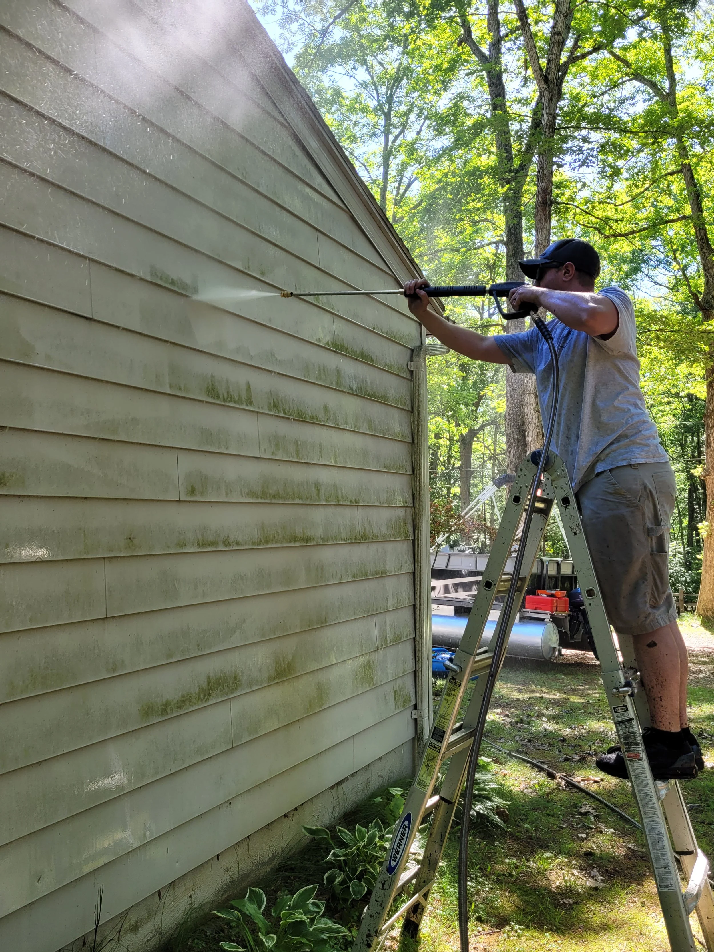 A person standing on a stepladder pressure washing the side of a house with green trees in the background.