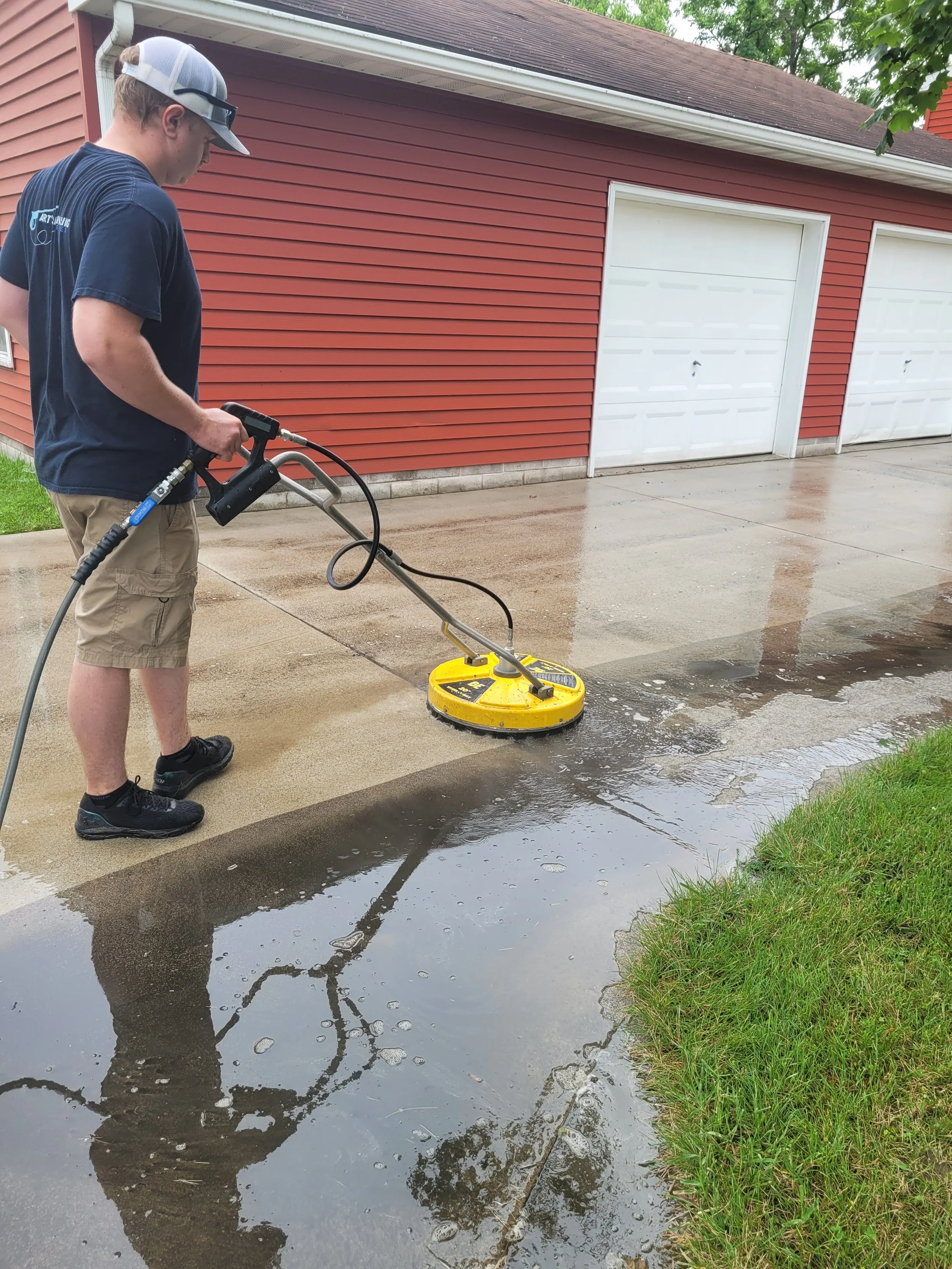 A person wearing a baseball cap, sunglasses, a navy T-shirt, and tan shorts is power washing a concrete driveway near a residential garage with a red house and a white garage door. The driveway has wet and dry areas, with water pooling on the surface