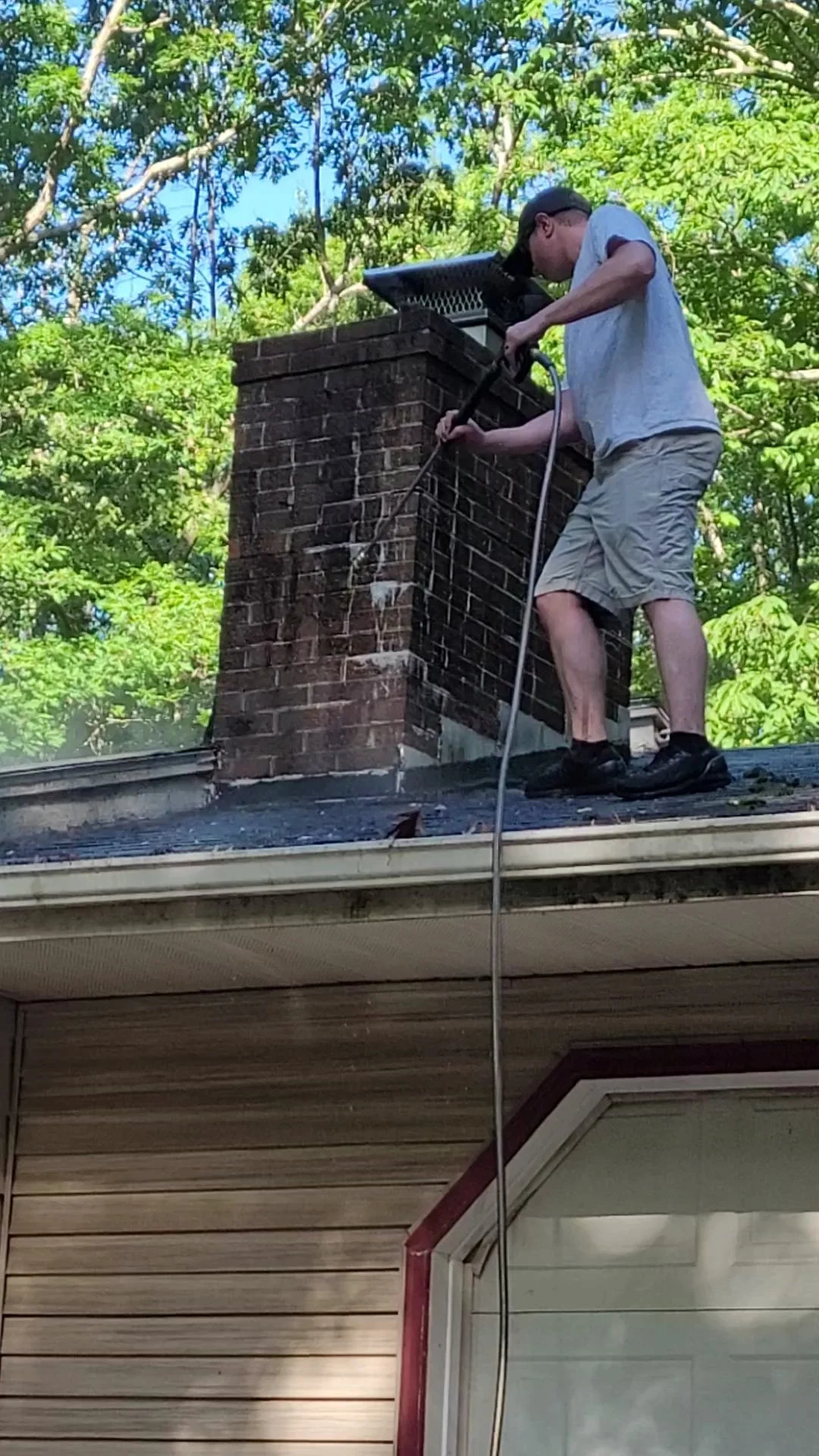 A man cleaning a chimney on the roof of a house surrounded by green trees.