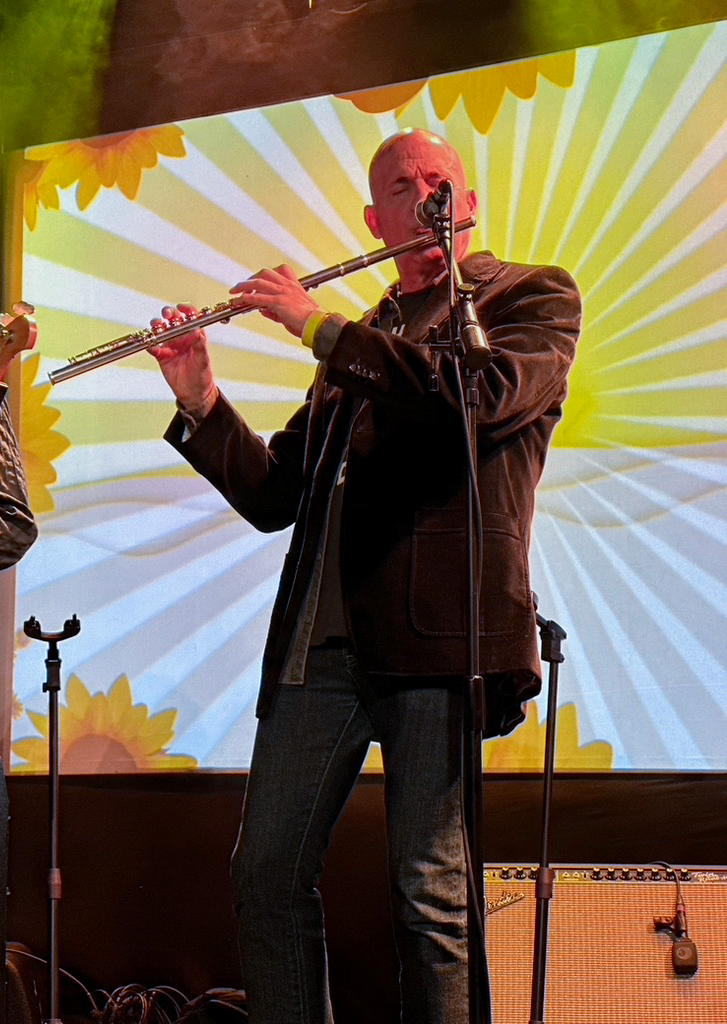 A man playing the flute on stage with a colorful sunflower and sunburst background.