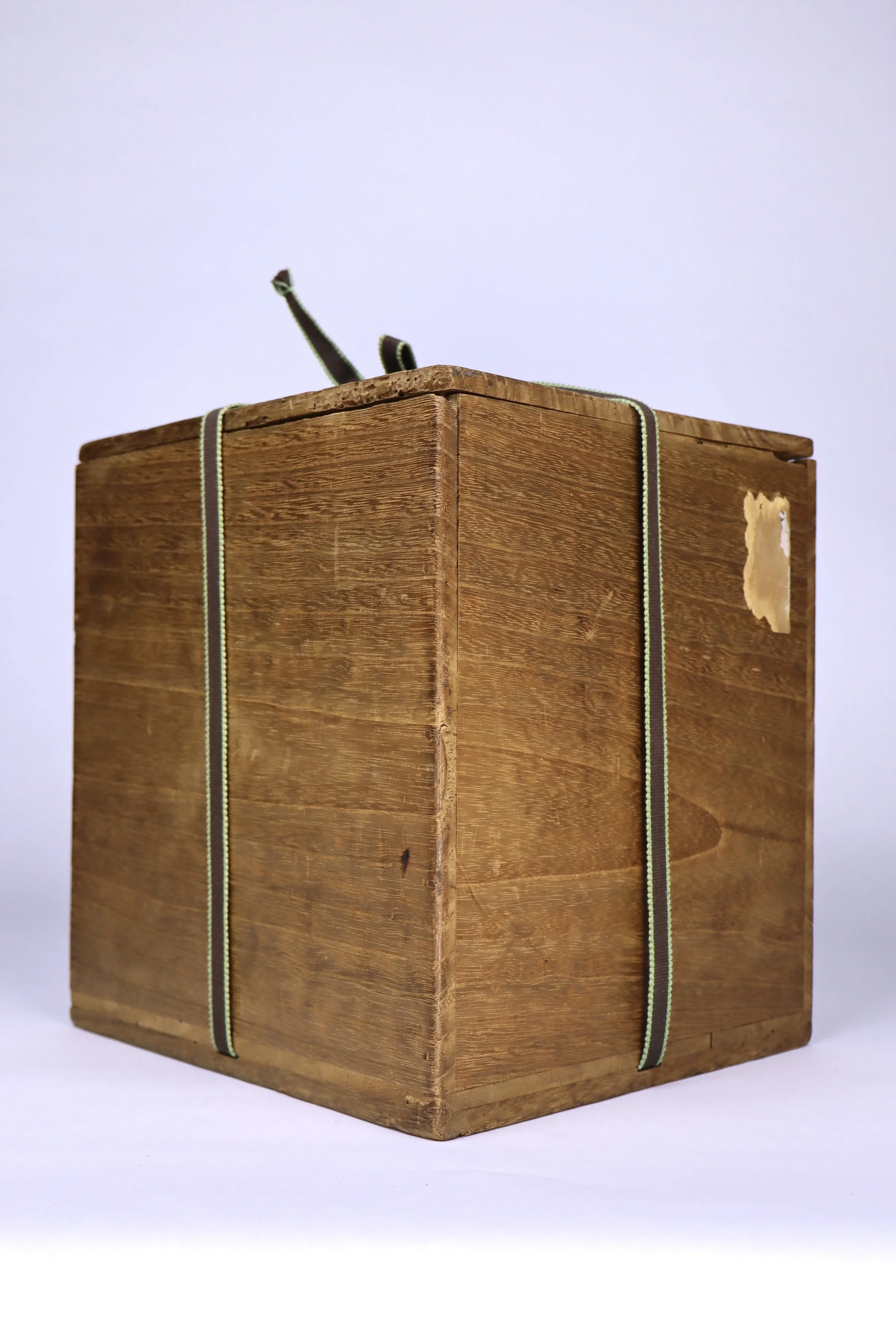 A wooden box with a corner visible, strapped with guitar straps, with a small patch of peeling paper on the side, placed on a white surface against a light background.