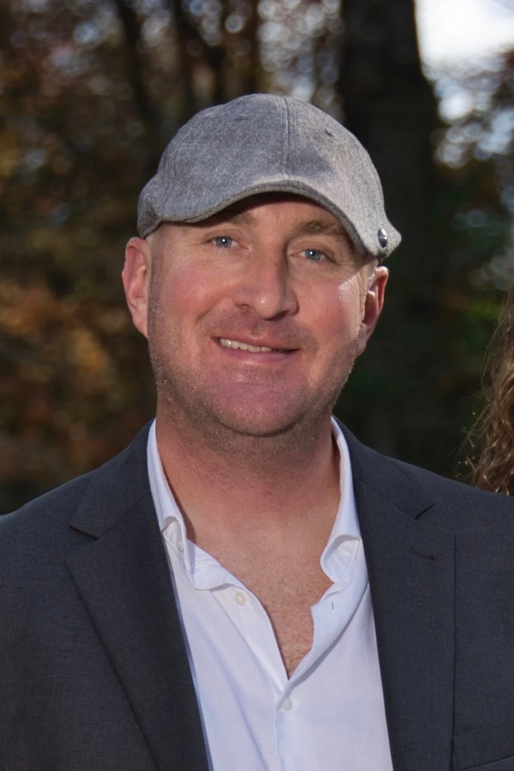A man smiling outdoors wearing a gray cap, a white shirt, and a dark blazer, with trees in the background.