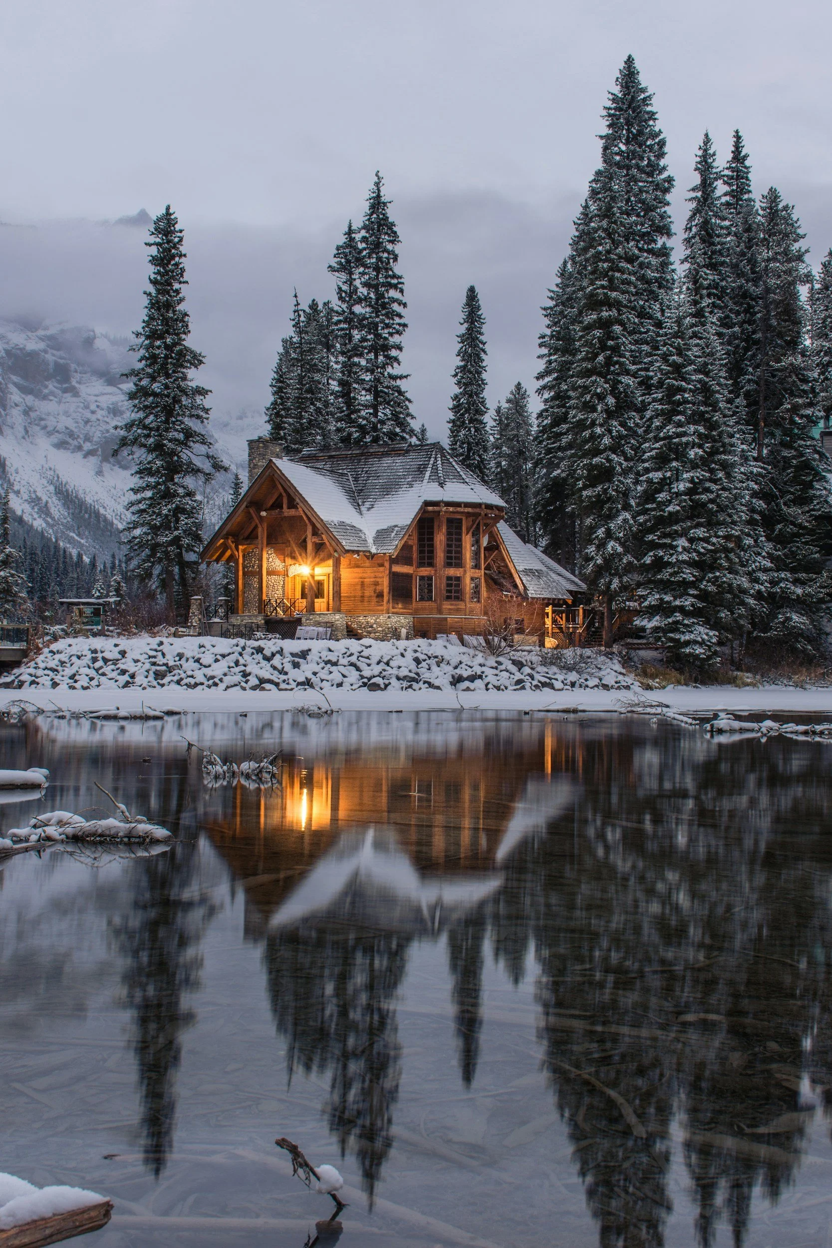 A wooden house with warm lights on at dusk, surrounded by snow-covered evergreen trees and mountains in the background, with a partially frozen river in the foreground.