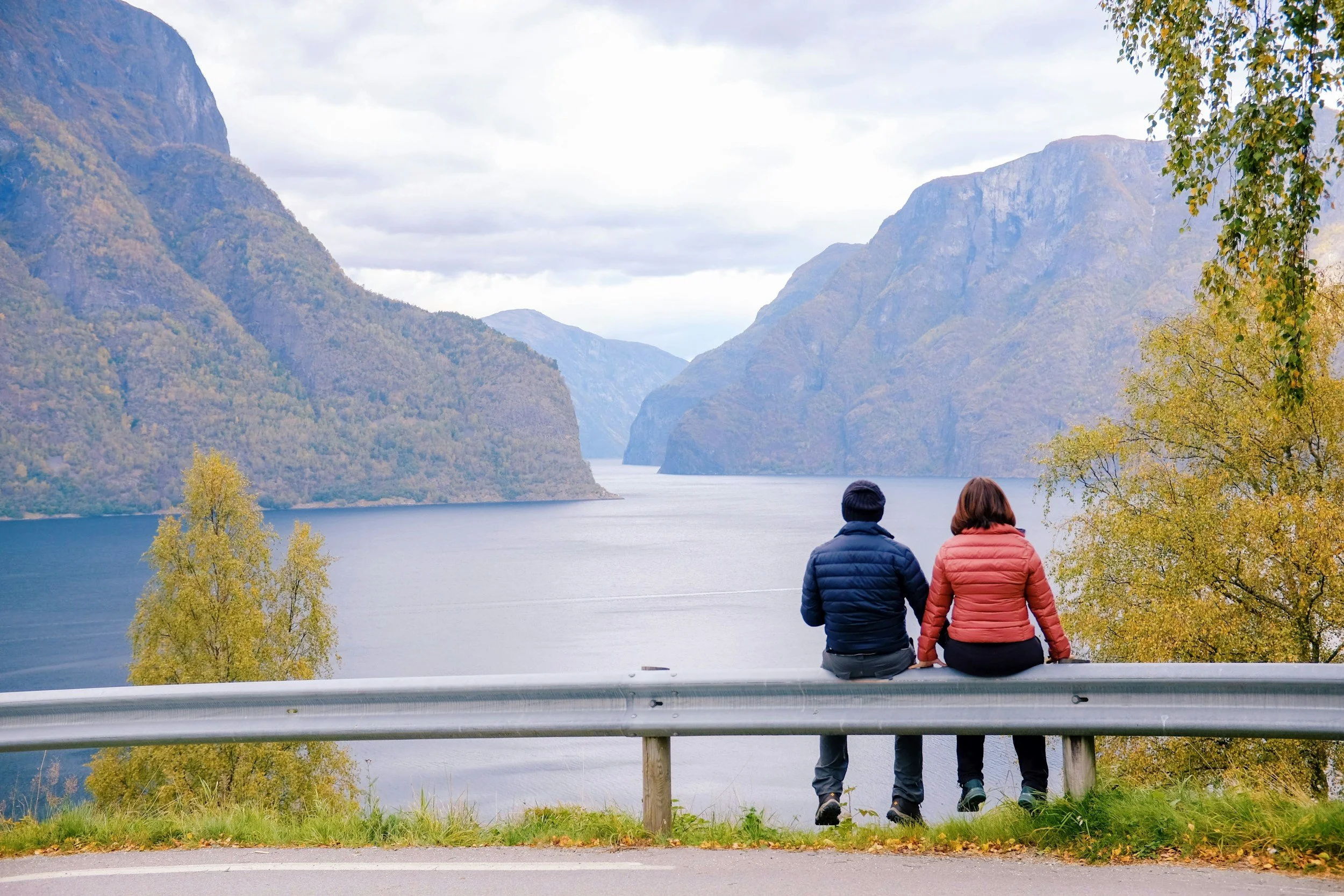 Two people sit on a guardrail overlooking a scenic lake surrounded by mountains, with trees showing fall colors.
