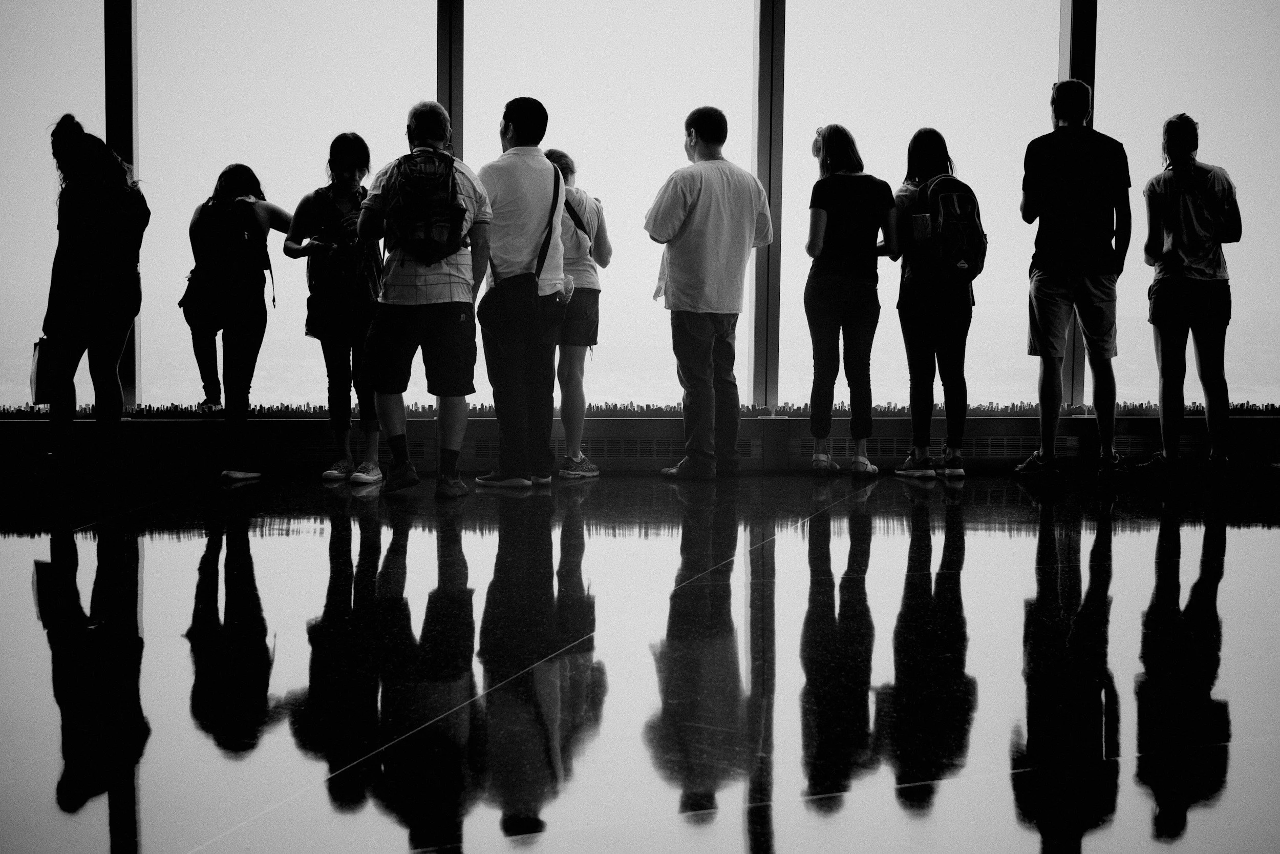 Silhouettes of a group of people standing in front of large windows, with their reflections visible on the shiny floor inside a modern building.