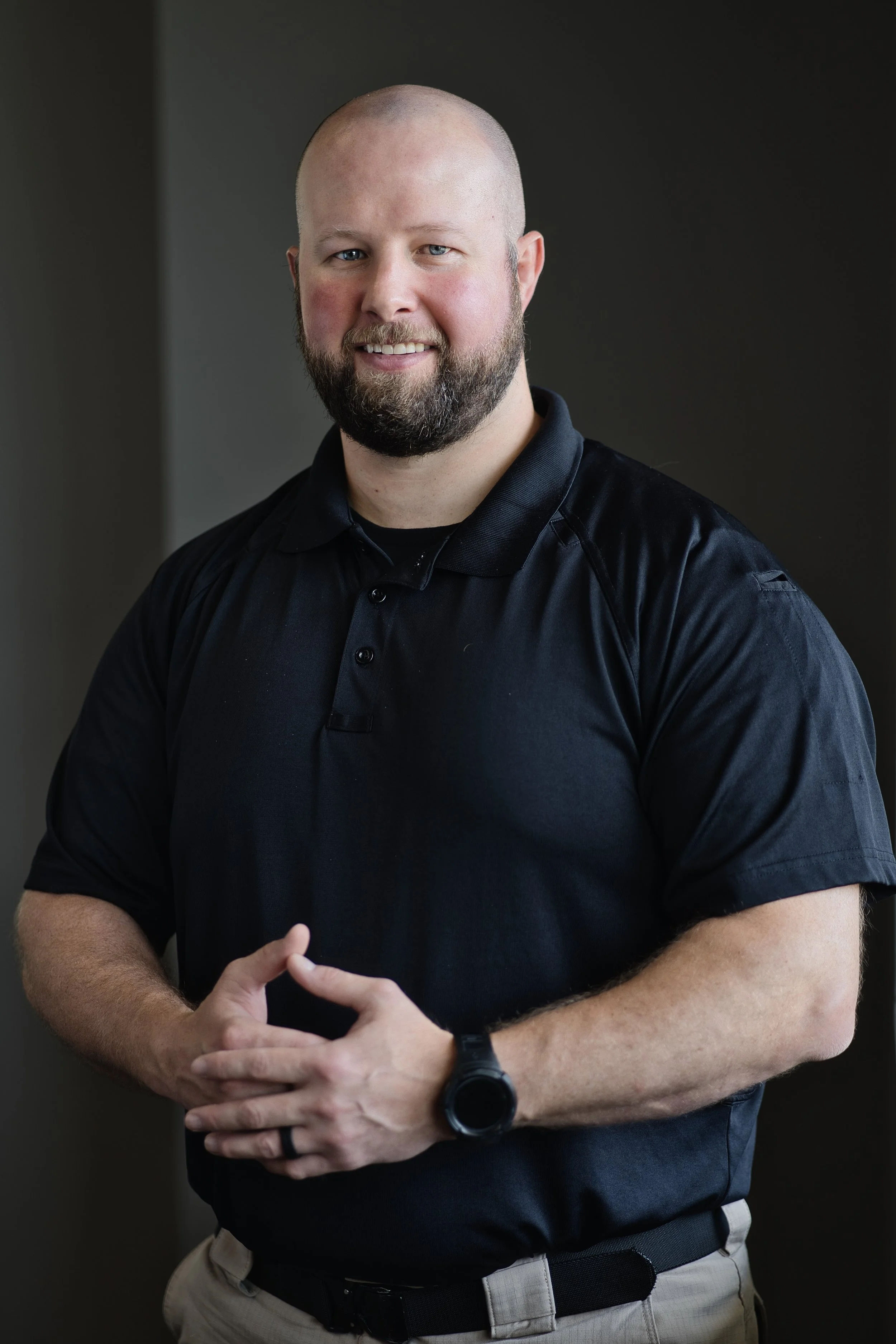A man with a beard and blue eyes, wearing a black polo shirt, beige pants, and a black watch, standing with arms crossed and smiling in front of a dark gray background.
