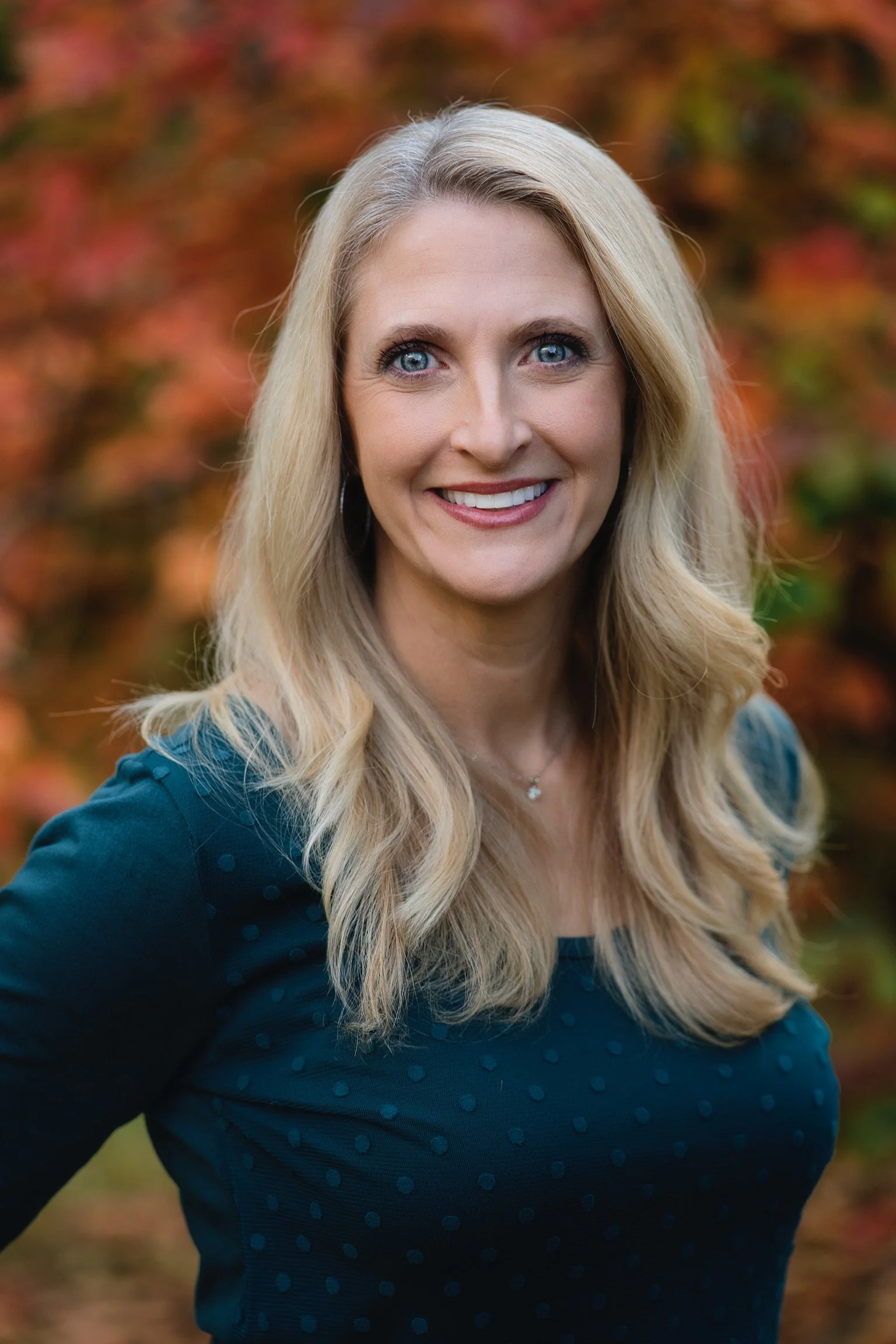 A woman with long blonde hair and blue eyes, smiling, outdoors with autumn foliage in the background.