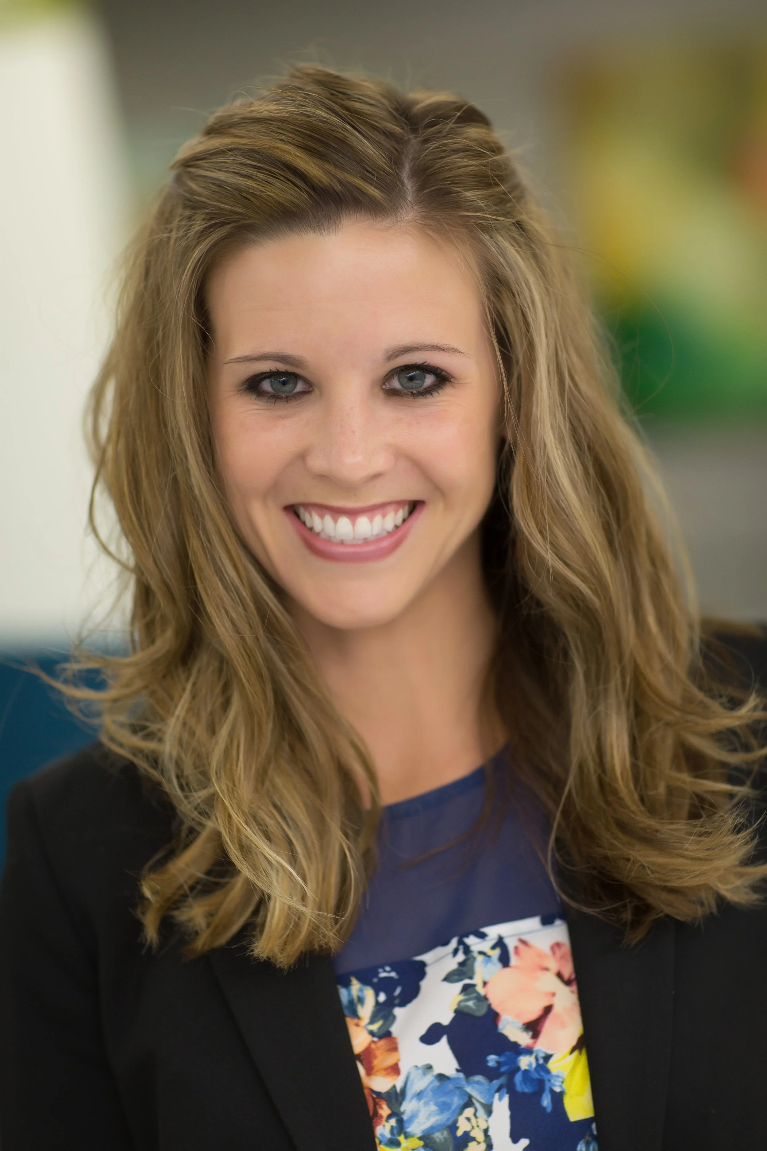 Close-up of a smiling young woman with long wavy blonde hair, wearing a floral blouse and a black blazer.