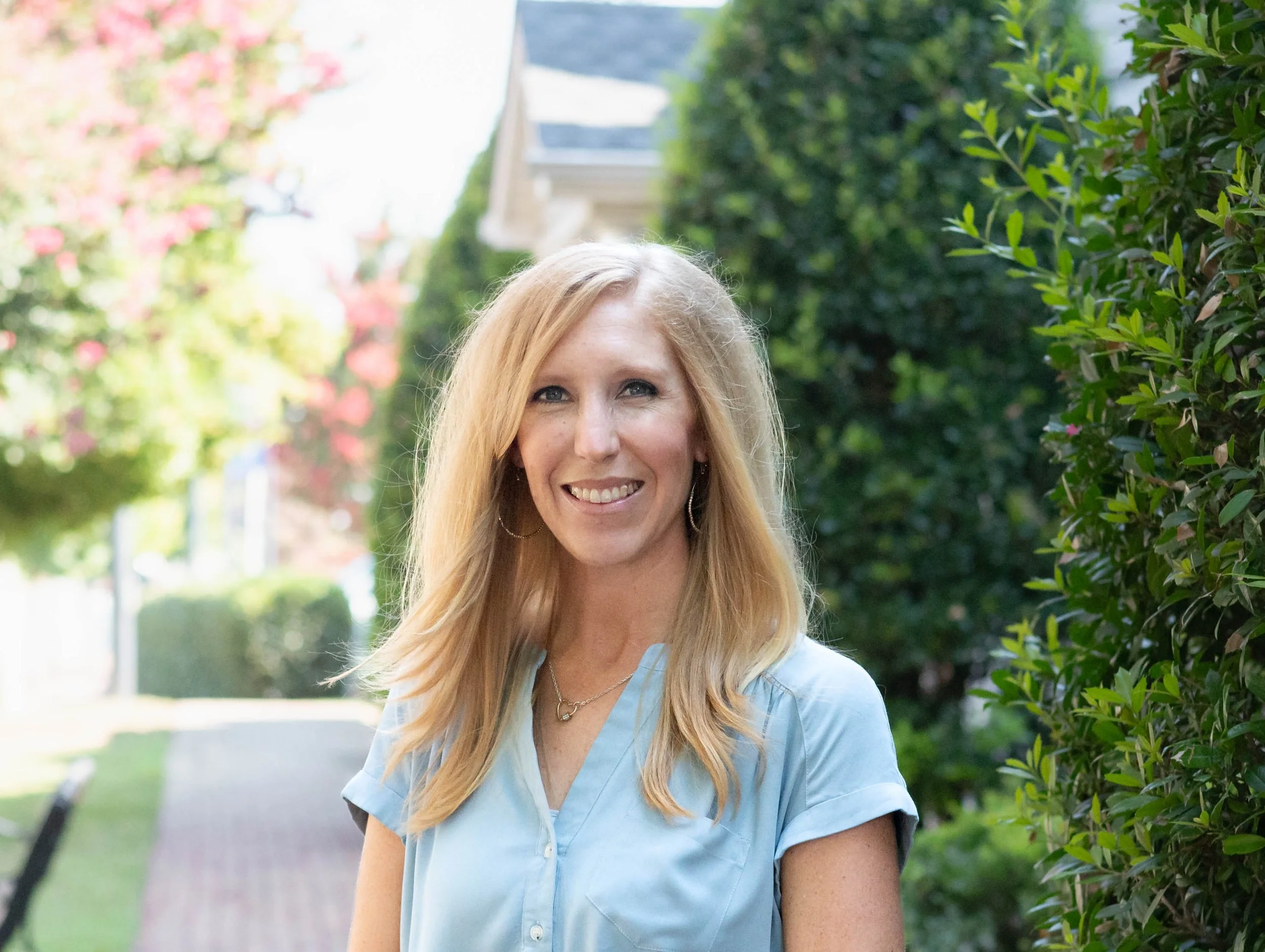 A smiling woman with long blonde hair wearing a light blue top standing outdoors beside a green hedge, with blurred trees and pathway in the background.