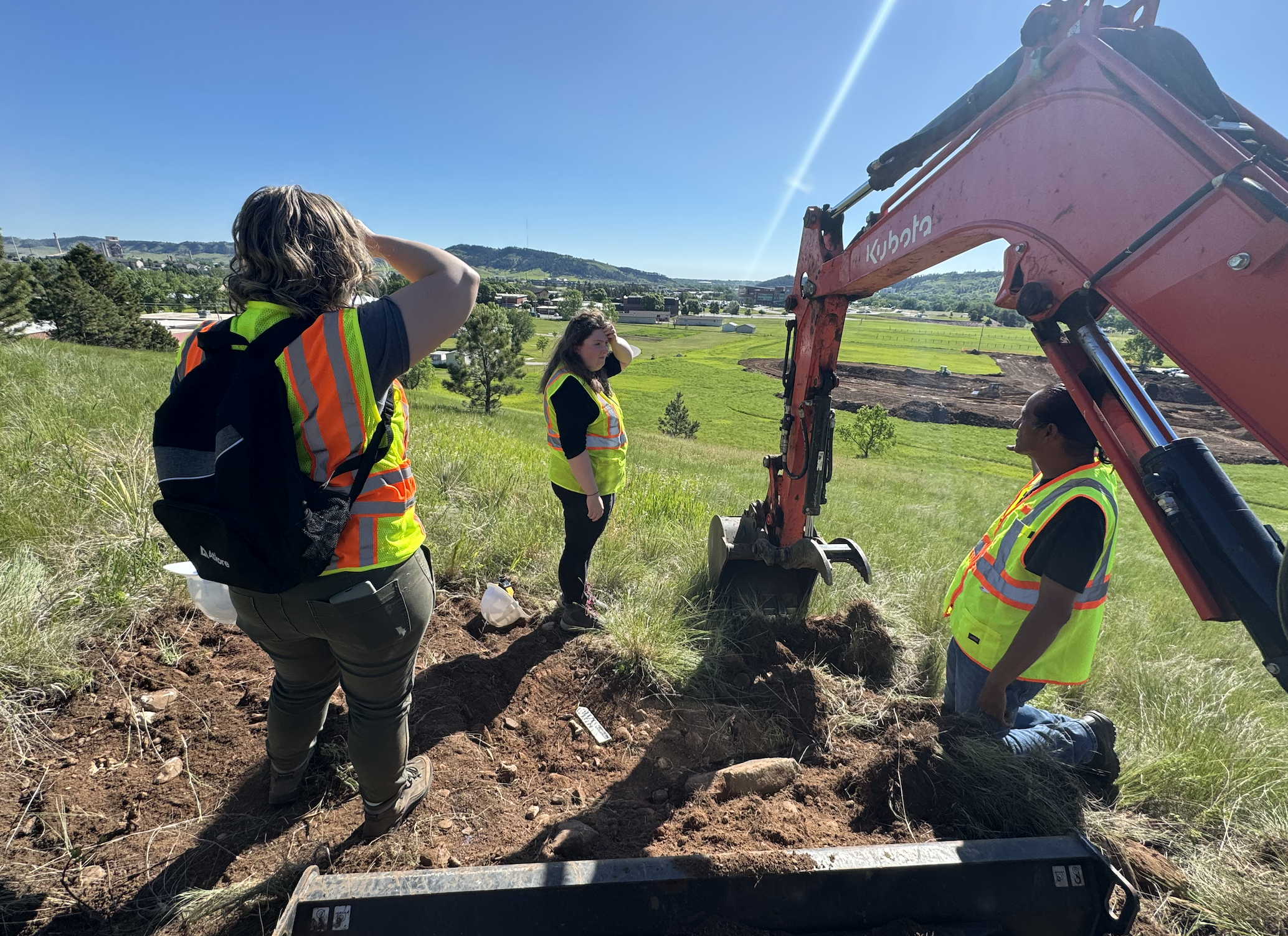 Three men planting a small plant or conducting a scientific experiment in a grassy hillside area with yellow wildflowers, some tools and planting trays on the ground.