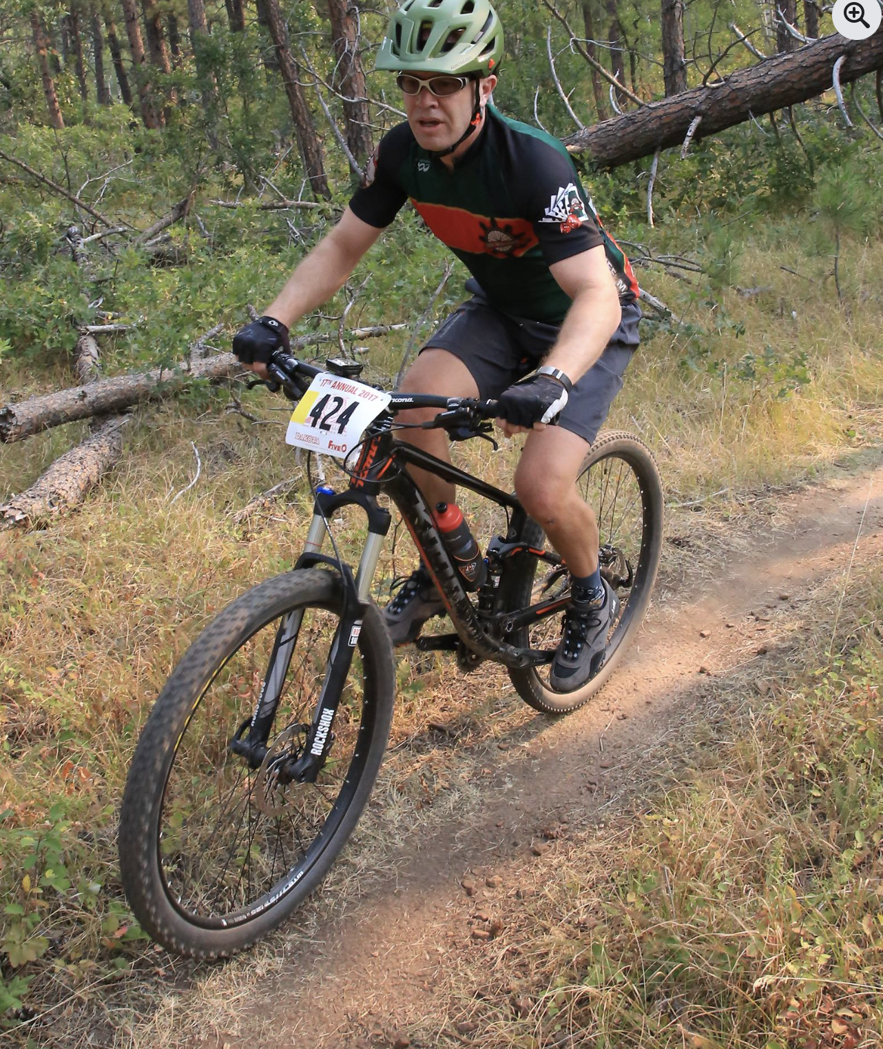 A man riding a mountain bike on a trail in a forest, wearing a green helmet, sunglasses, a black and green cycling jersey, gray shorts, and gloves, with a race number 424 attached to the bike.