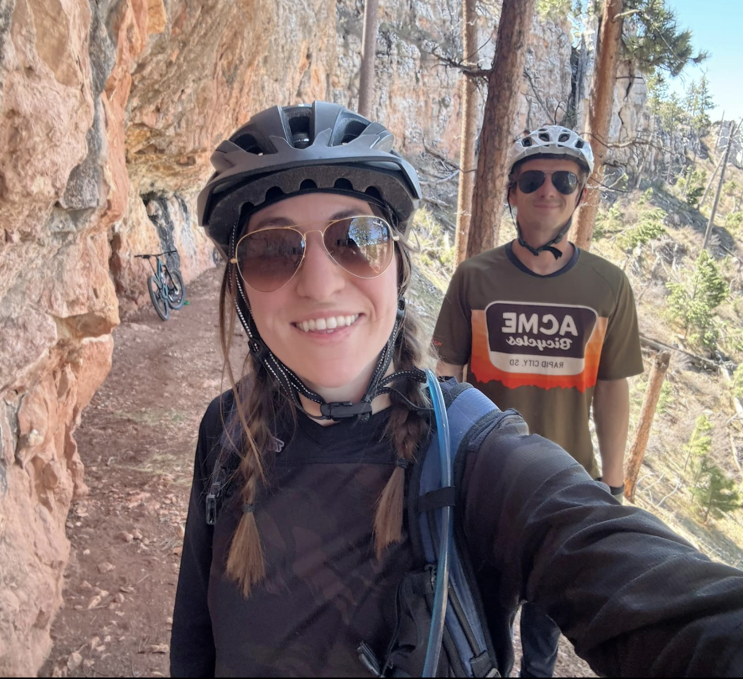 A smiling woman and a man wearing bicycles helmets and sunglasses on a mountain trail surrounded by trees and rocky cliffs.