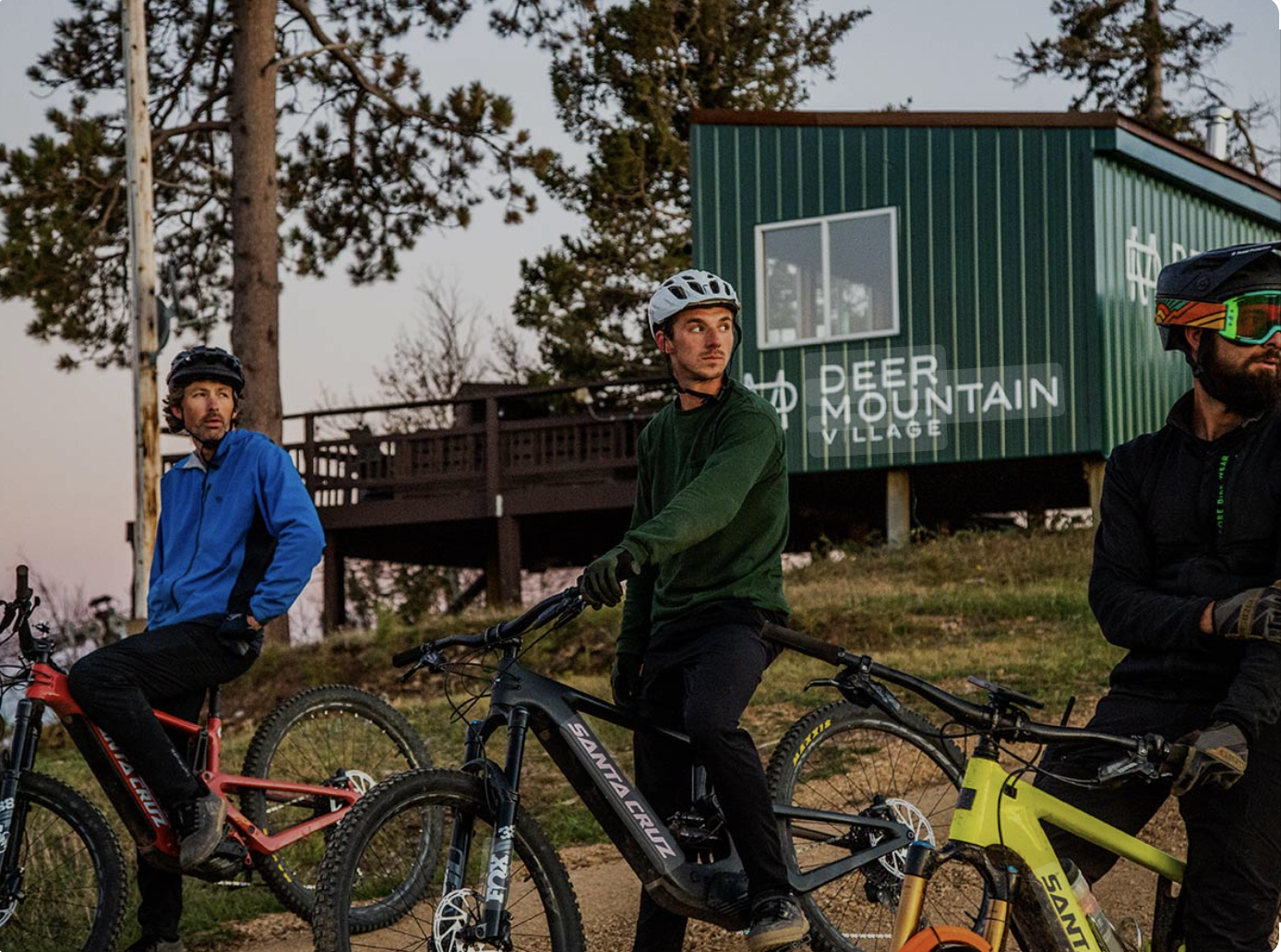 Three men with mountain bikes and helmets standing outdoors near a green building with a sign that reads "Deer Mountain Village", during dusk, with trees in the background.