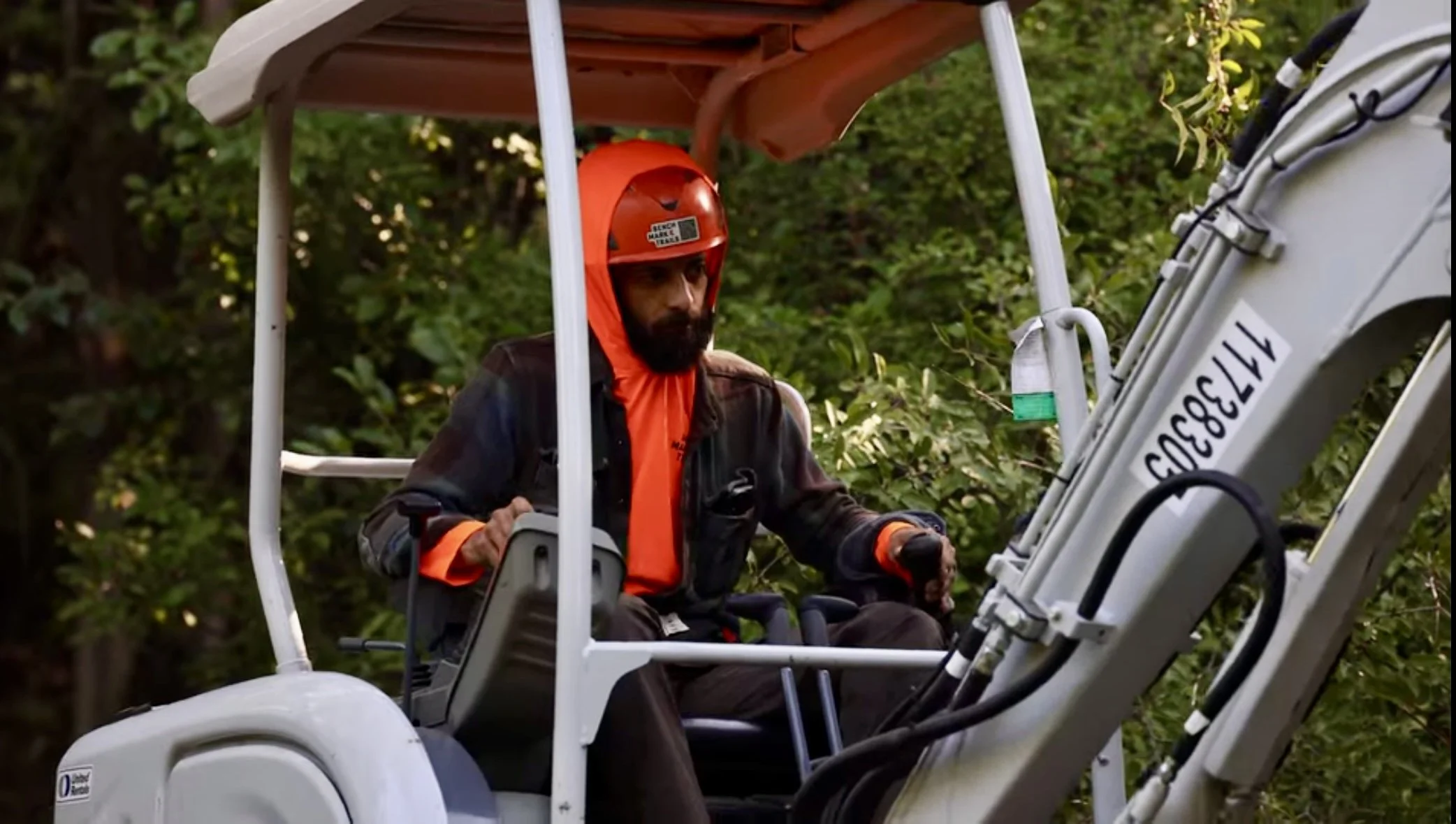 A man with a beard wearing a safety helmet and orange clothing operating a mini excavator in a wooded area. MTB Trailbuilding. Singletrack Construction.