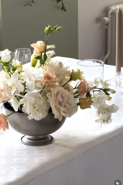 En blomsterarrangemang med vita, rosa och beige blommor i en metallskål på ett vitt dukat bord.