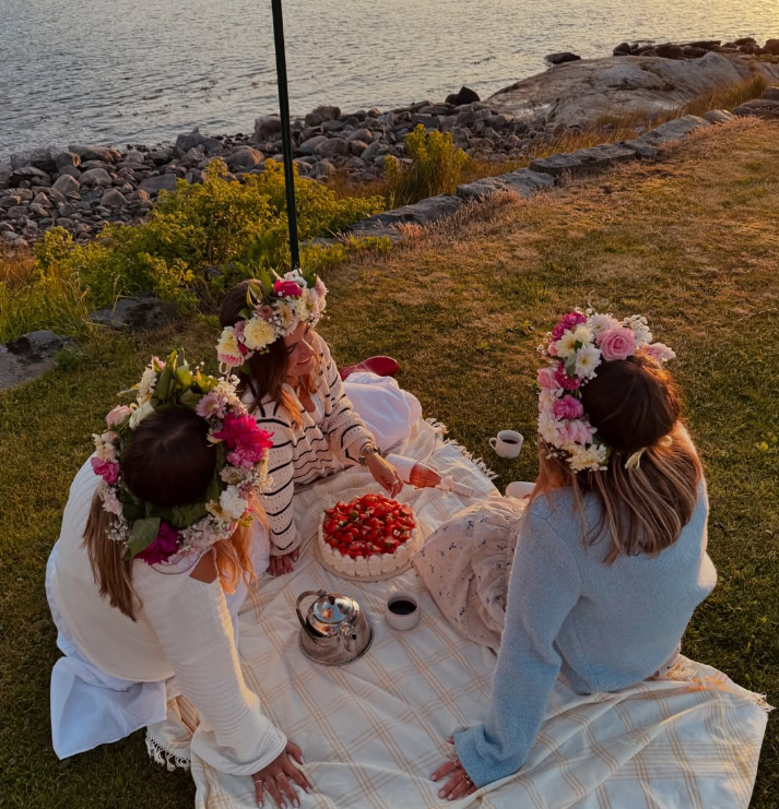 Tre kvinnor i blomsterkransar njuter av en picknick med jordgubbstårta och kaffe vid en sjö, under solnedgången.
