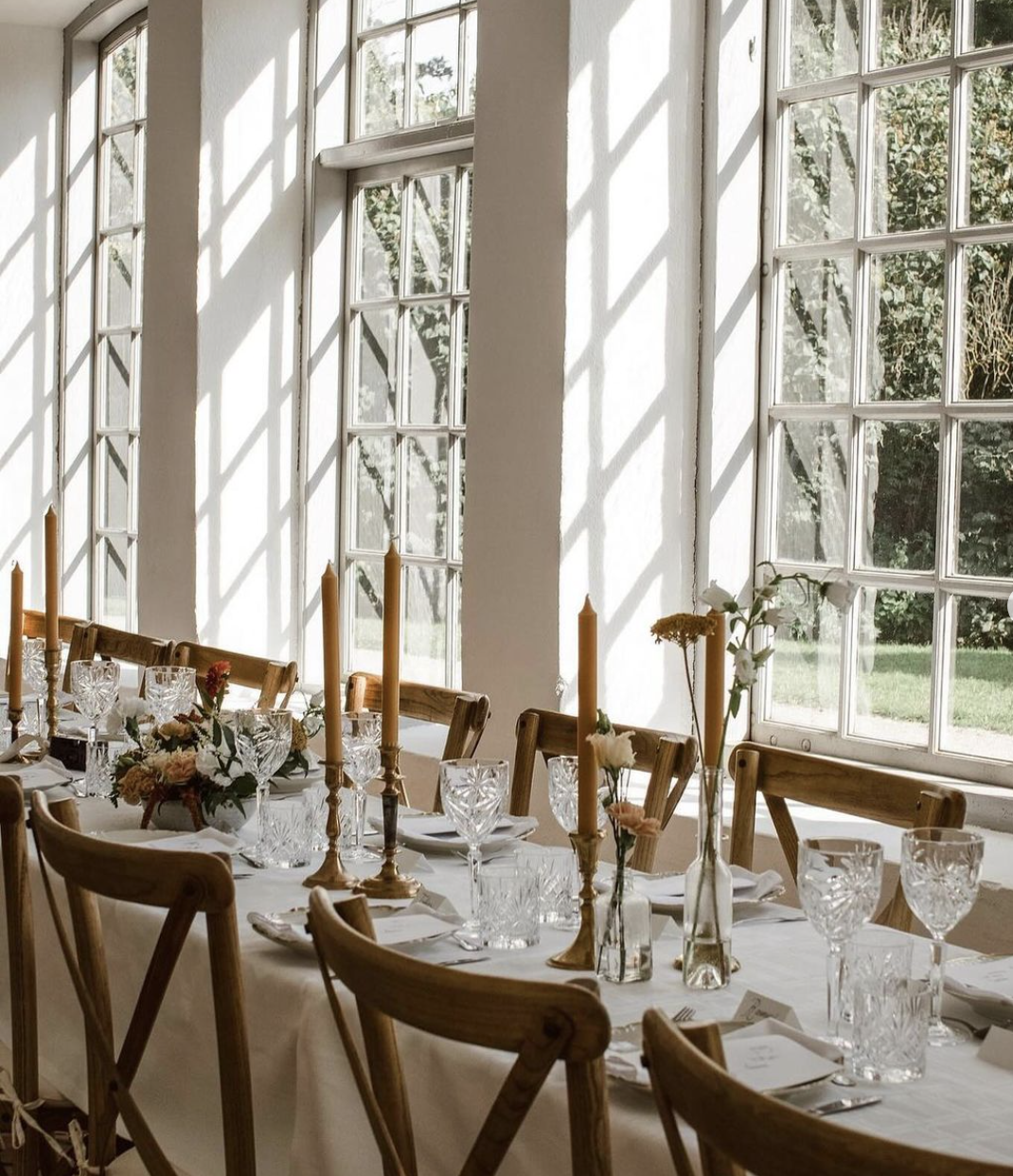 Elegant middagsdukning med glas, tallrikar, gafflar, knivar, ljusstakar och blommor i ett vårrumsdiningrummet med stora fönster och solljus.