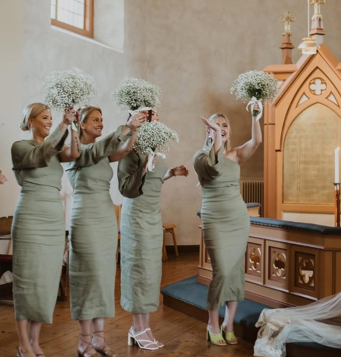 Sex kvinnor i brudklänningar och kavajer håller buketter av små vita blommor i en kyrka, förmodligen vid ett bröllop.