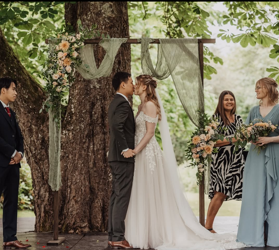 En brud och en brudgum kysser varandra under ett träd vid deras utomhusbröllop, omgivet av blommor och vackra gardiner, med två kvinnor som ser på och ler.