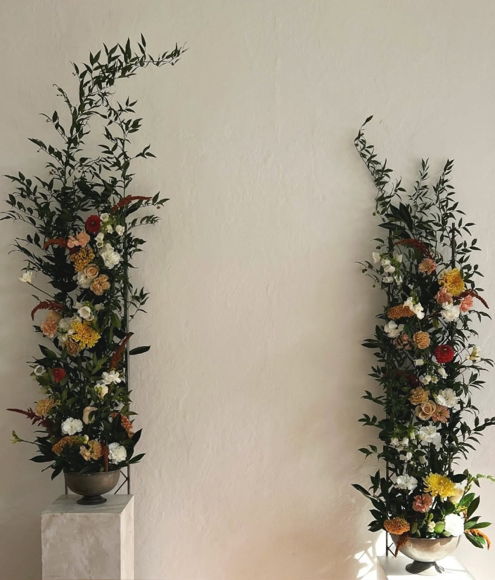 Två blomsterarrangemang i vasader, placerade på vita pelare mot en beige vägg.