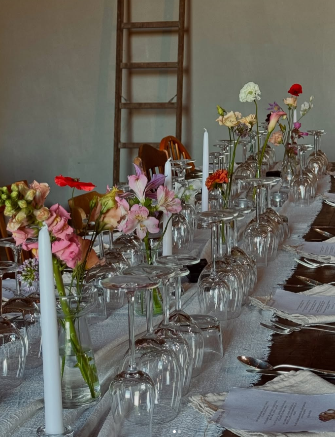 En lång bordssättningen med avslutade glas, blommor i vaser, vita ljus och servetter i ett inomhusrum.