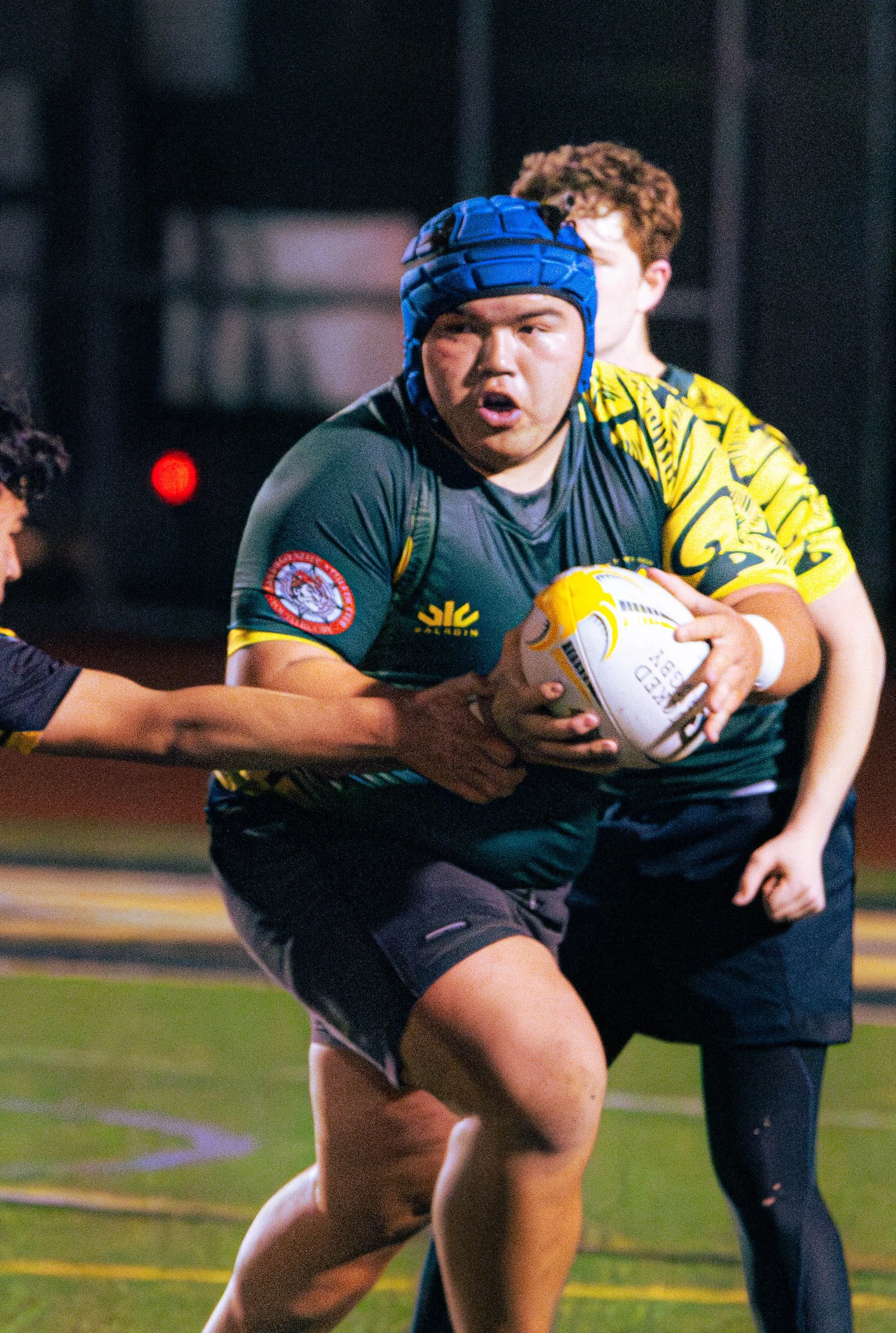 A rugby player holding a rugby ball, wearing a dark green and yellow jersey, a blue headgear, and black shorts, on a field during a game.