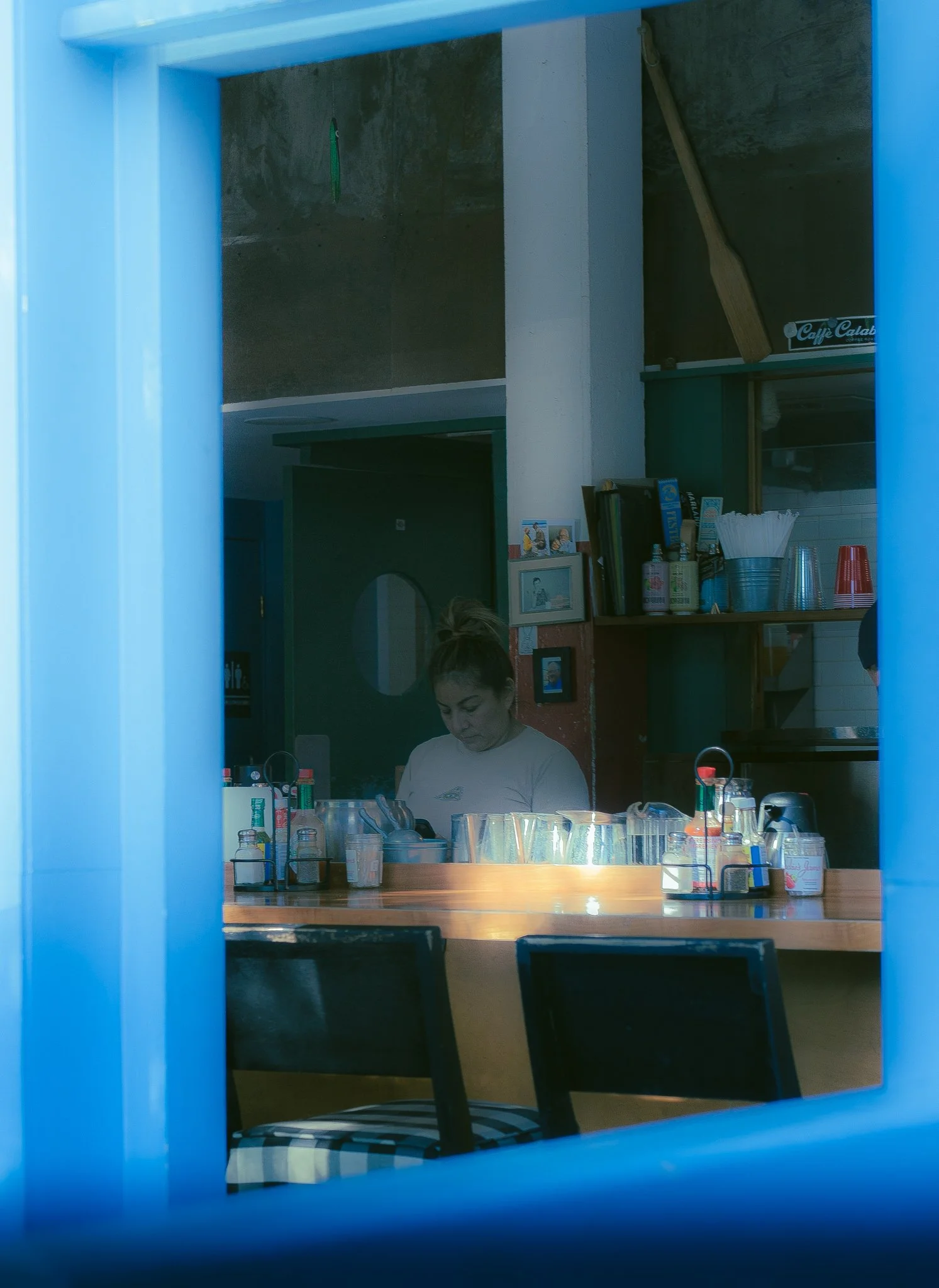 A woman preparing food in a restaurant kitchen, viewed through a blue window frame.