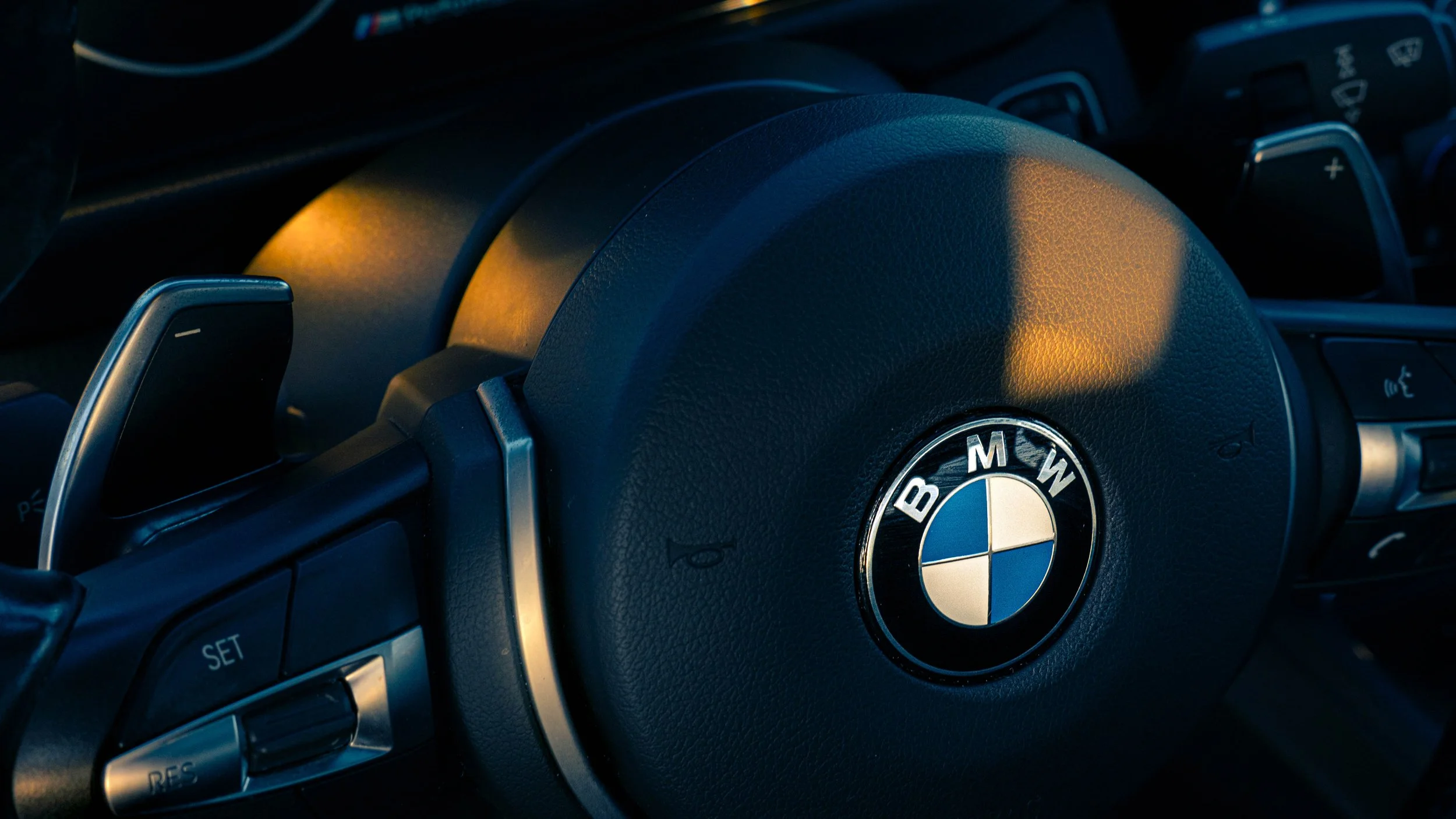 Close-up of a BMW steering wheel with the company's logo in the center, illuminated by warm light, part of the dashboard with control buttons visible.