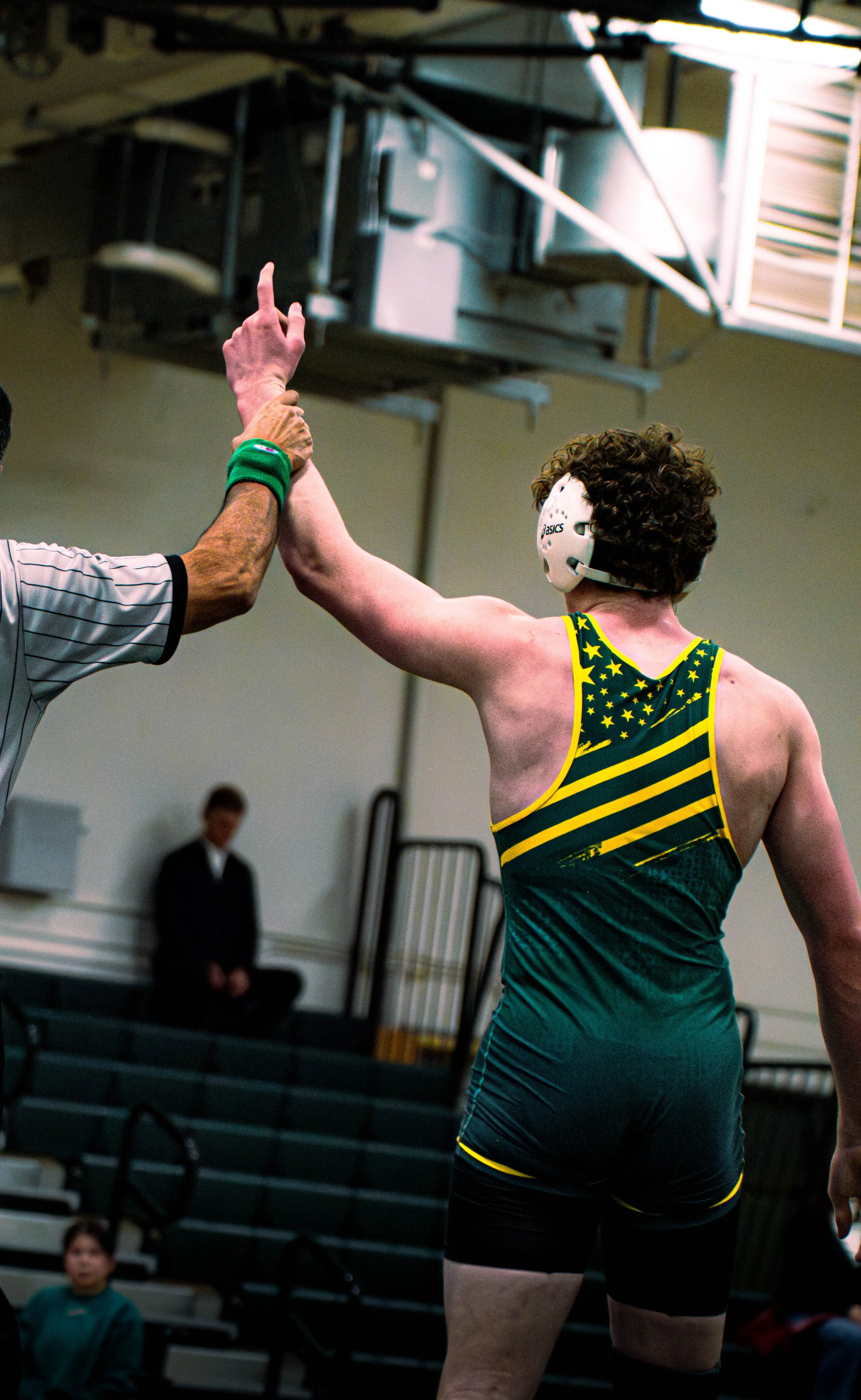 A wrestler in a yellow and green singlet receives a high five from a referee during a match, with a referee and audience members in the background.