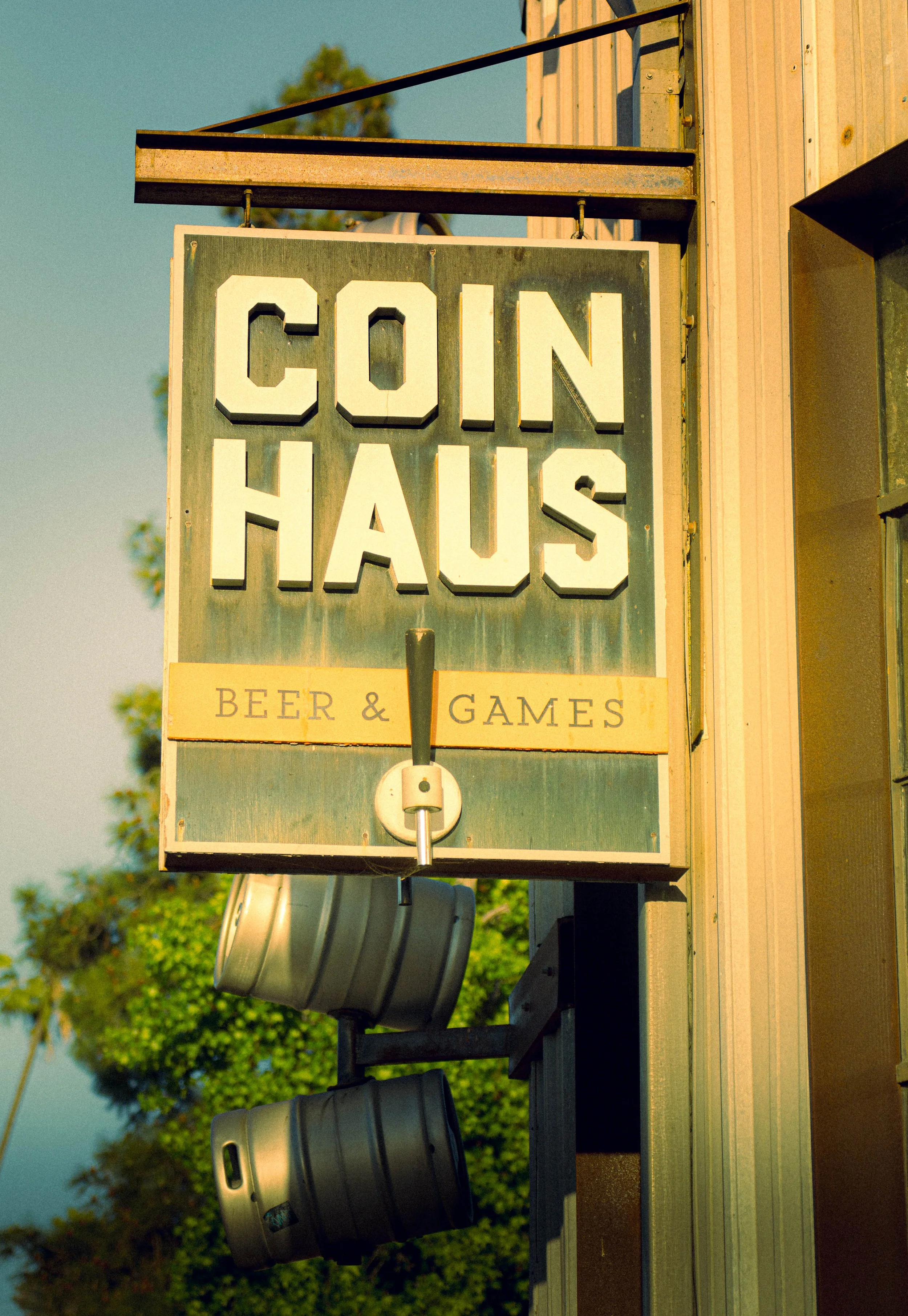 A sign for a business called 'Coin Haus' that offers beer and games, with a dark-colored mailbox or trash can below the sign, attached to a building exterior.