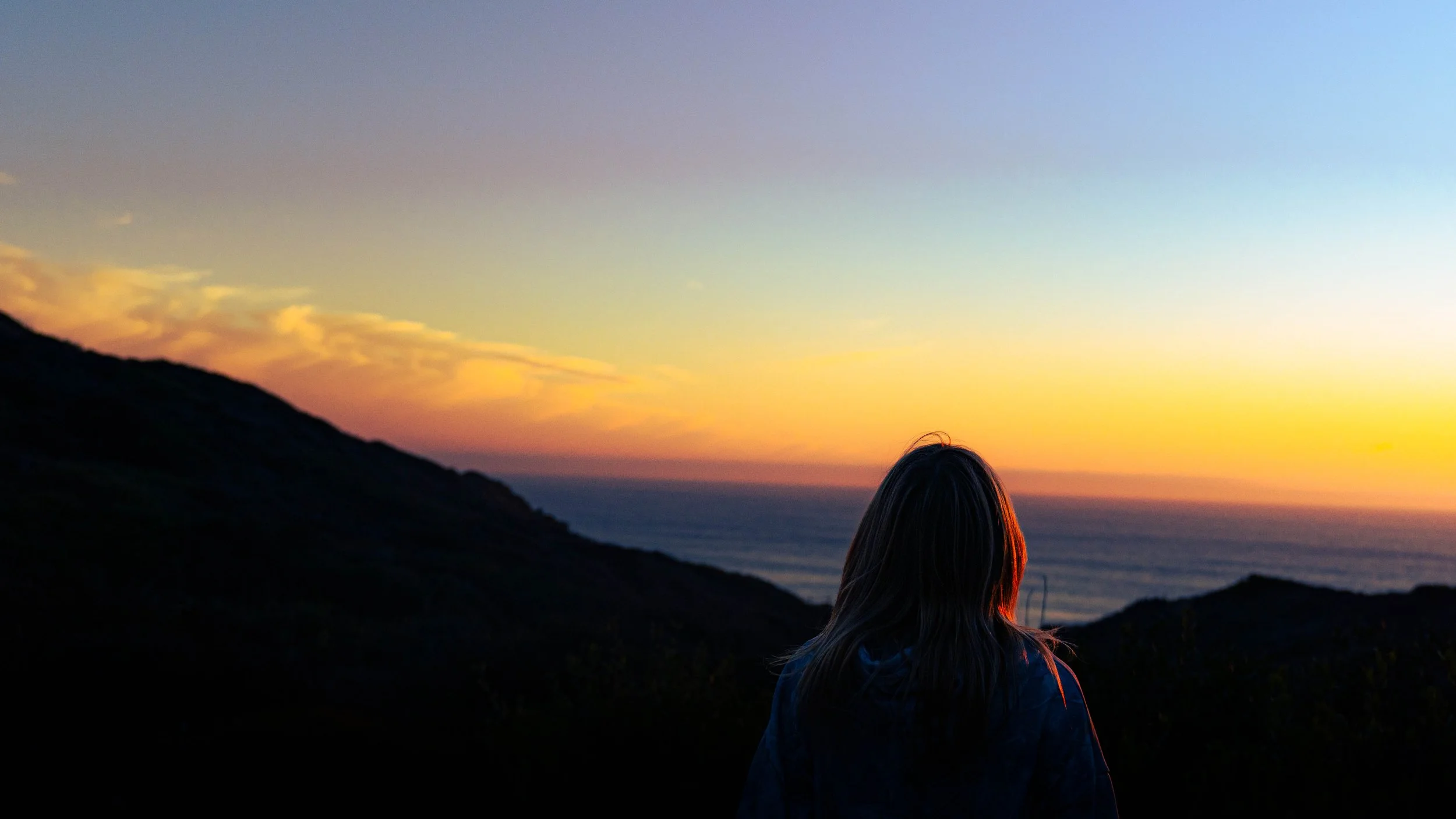 A person with long hair, wearing a jacket, sitting outdoors on a hillside during sunset, overlooking the ocean and colorful sky.
