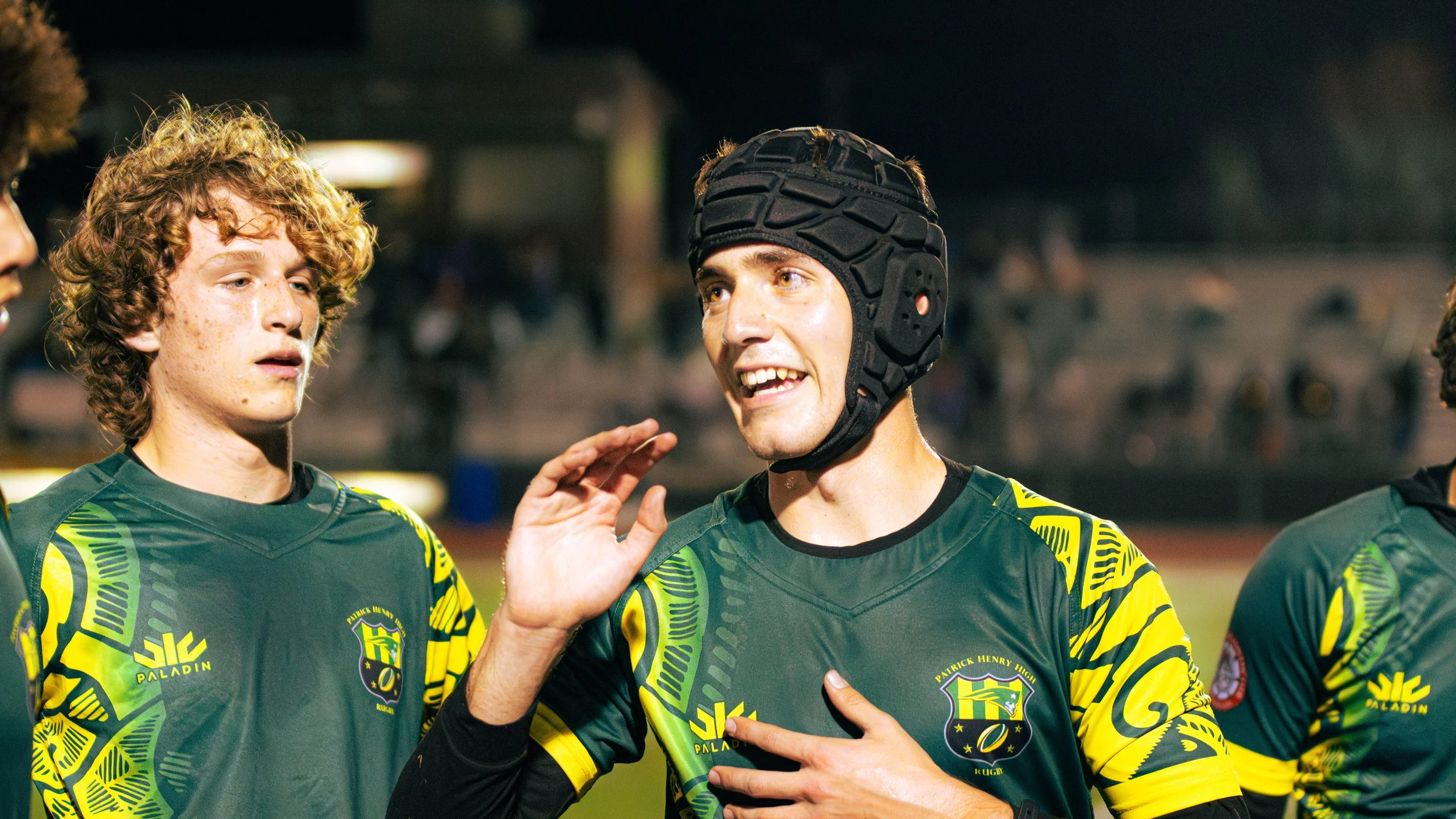 Rugby players in green and yellow jerseys, one wearing a black helmet, talking during a game at night.