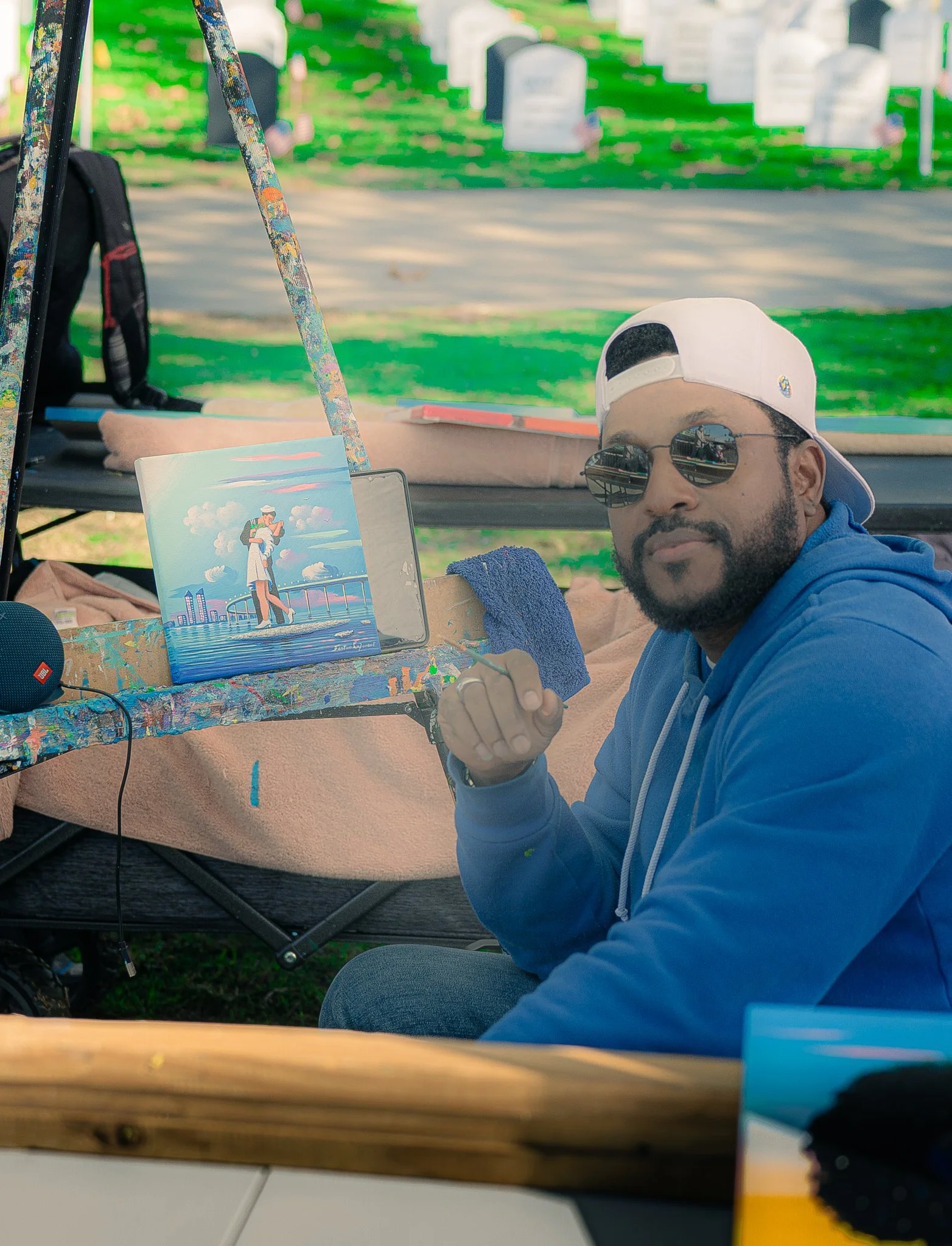A man wearing a white cap backwards, sunglasses, and a gray hoodie sitting on a bench with a colorful art piece in front of him at an outdoor location. Graves and tombstones are visible in the background.